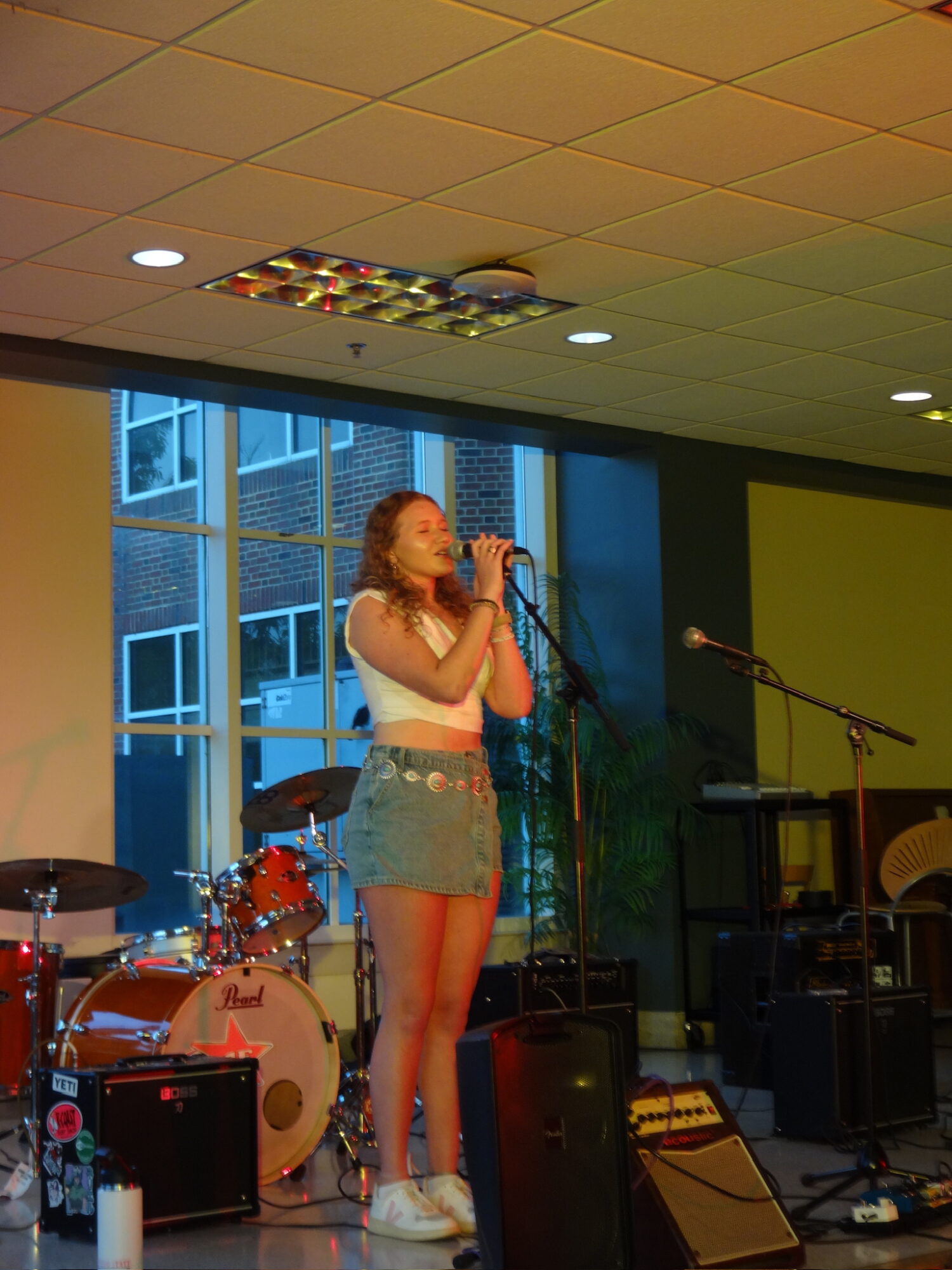Young woman singing into a microphone on stage with musical instruments and large windows behind her.