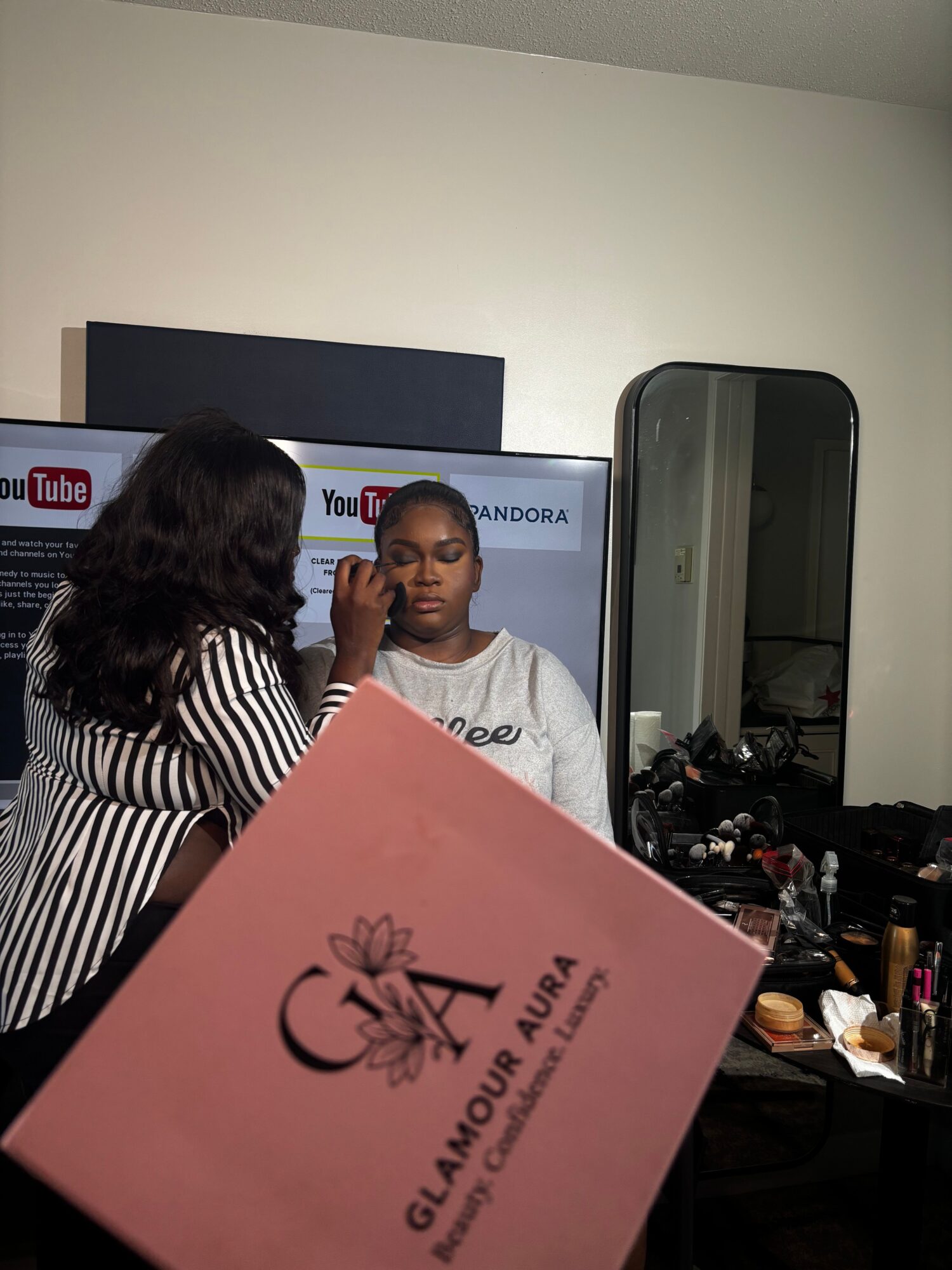 Two women in a room with a mirror and a TV screen, one holding a pink box with black text, facing each other.
