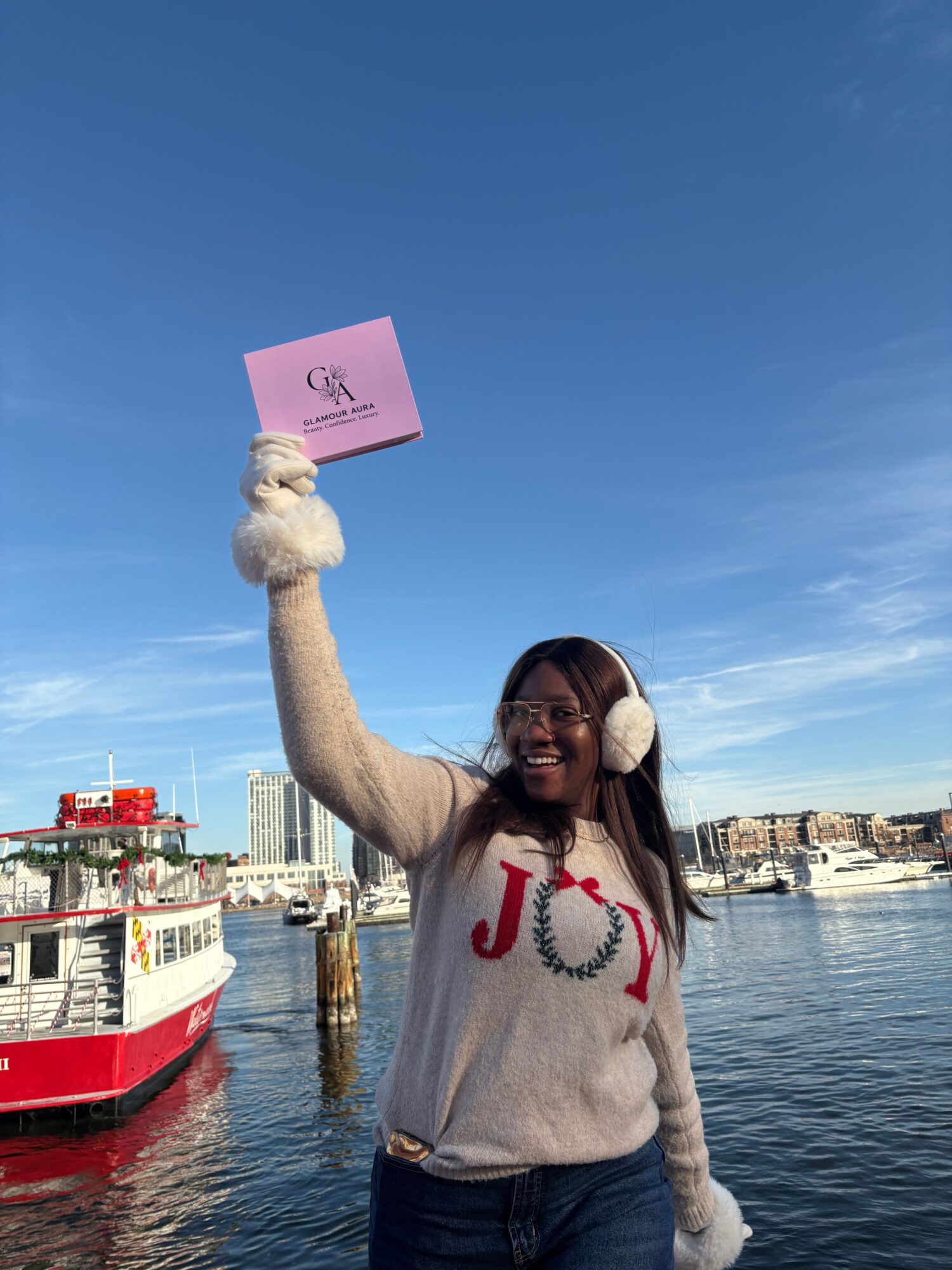 Person standing by water holding a pink sign, wearing a gray hoodie with red text, with boats and buildings in the background.