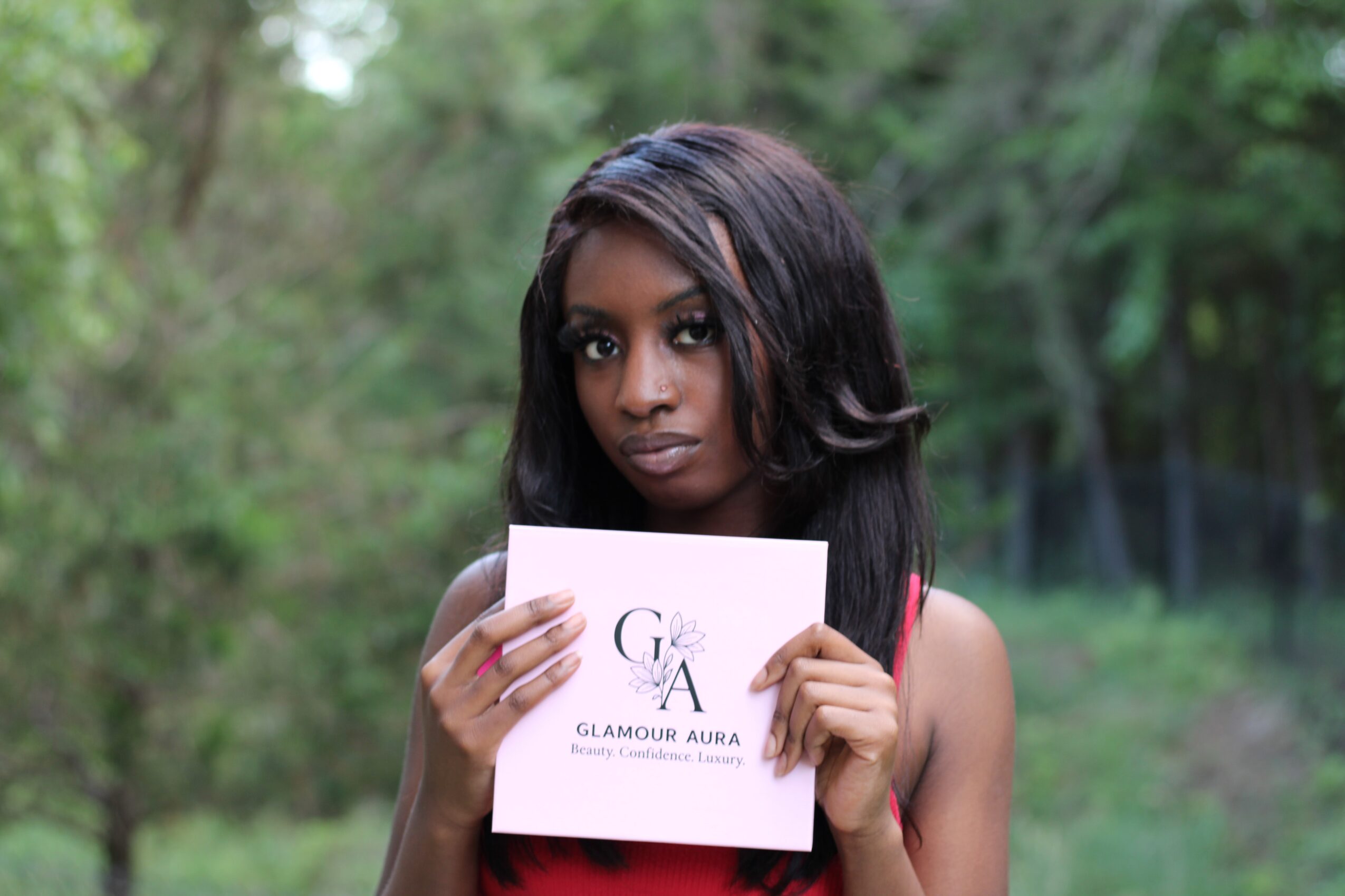 Young woman with dark hair holding a sign outdoors, surrounded by greenery.