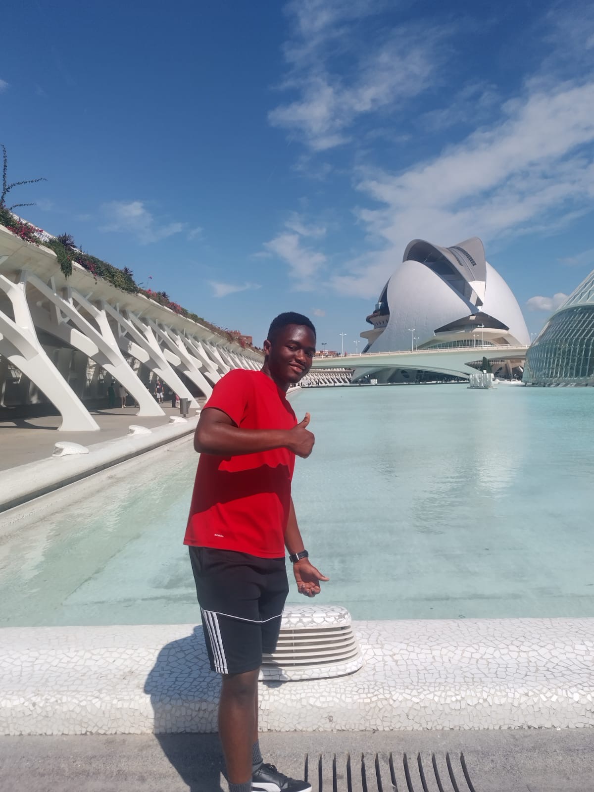 Young man in red shirt and black shorts standing near water with modern building and blue sky in background.