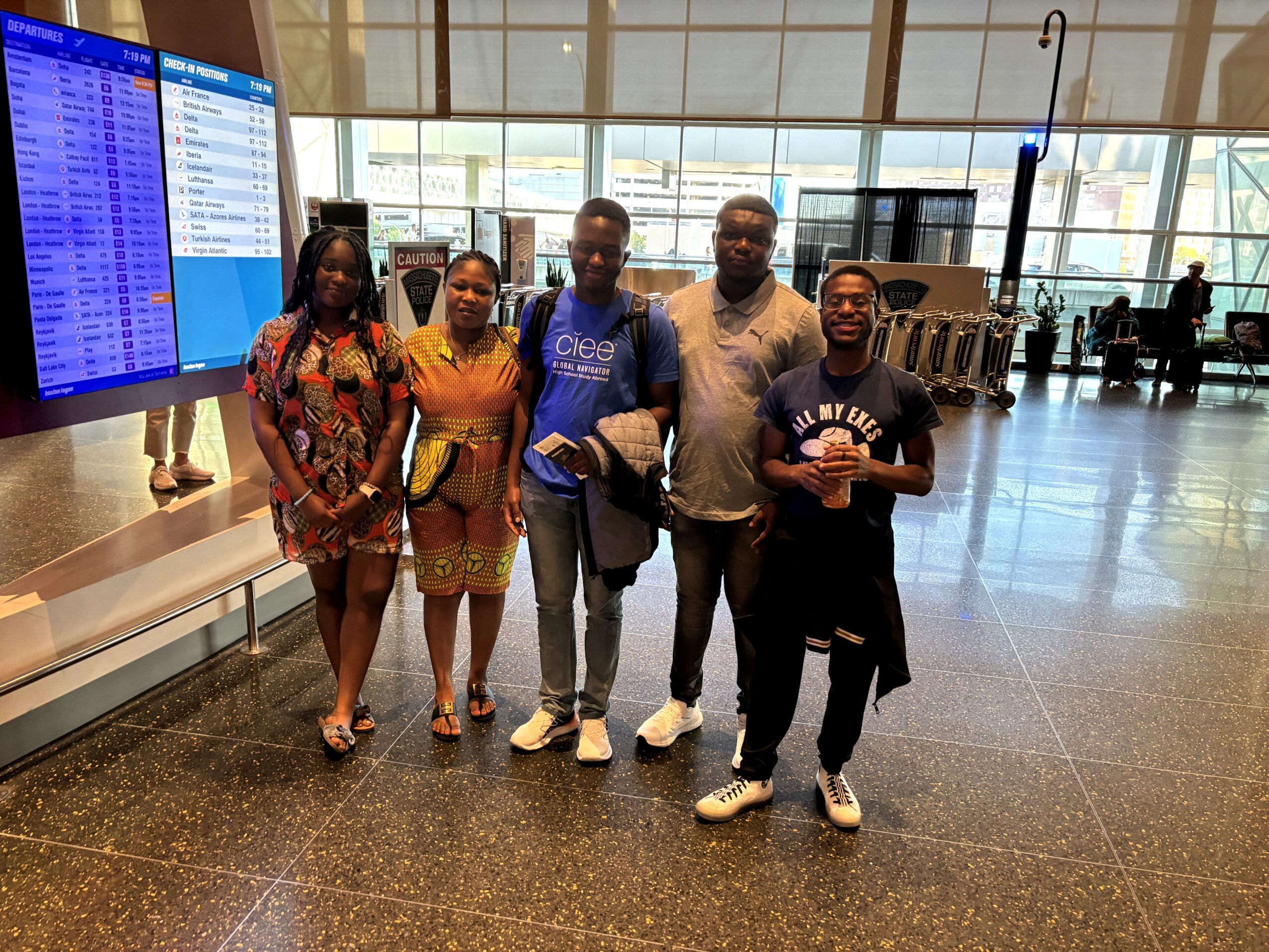 Five people standing in an airport terminal, with large windows and luggage carts in the background.