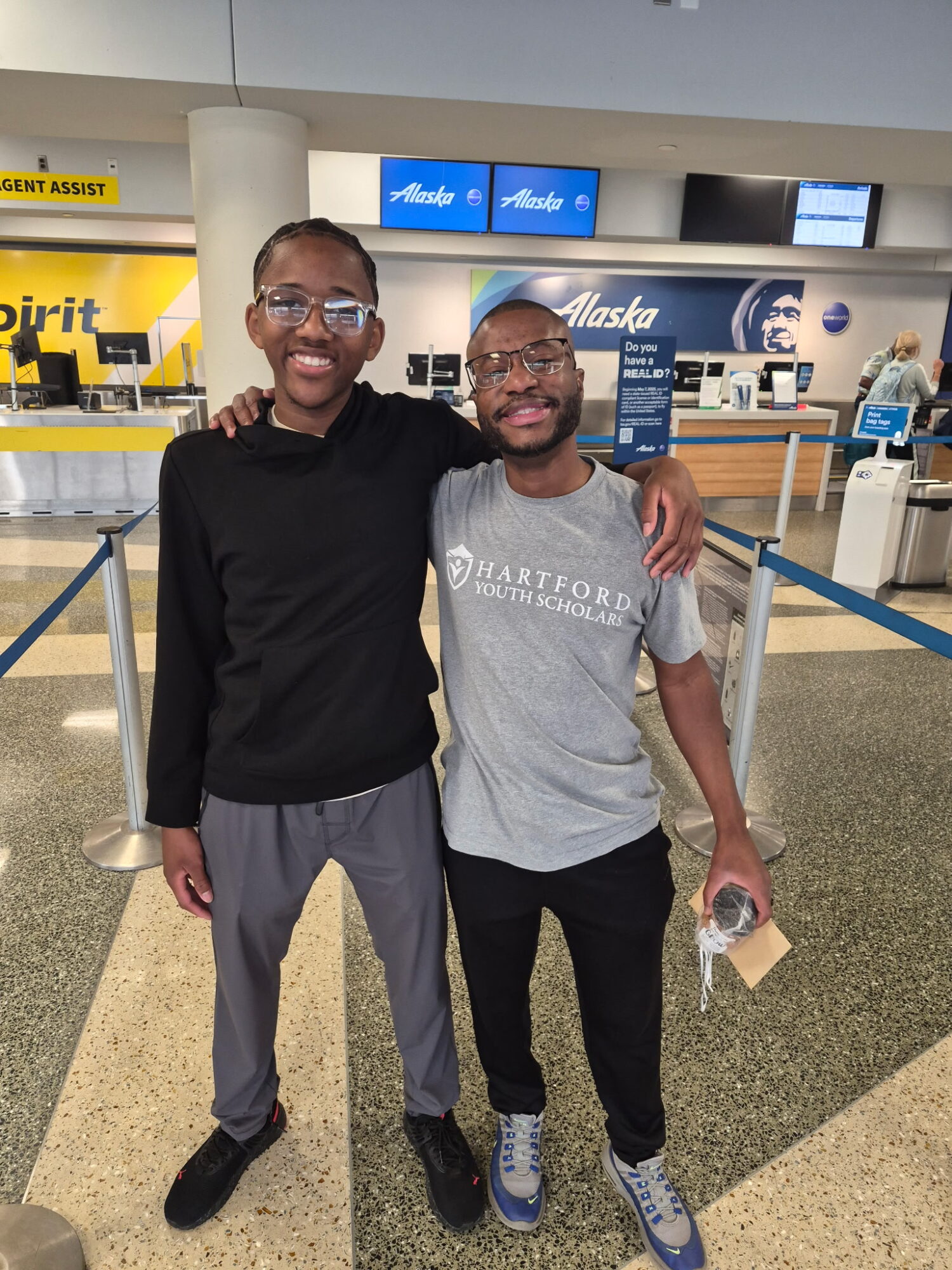 Two men stand together in an airport, smiling, with one arm around each other's shoulders, behind a metal barrier.
