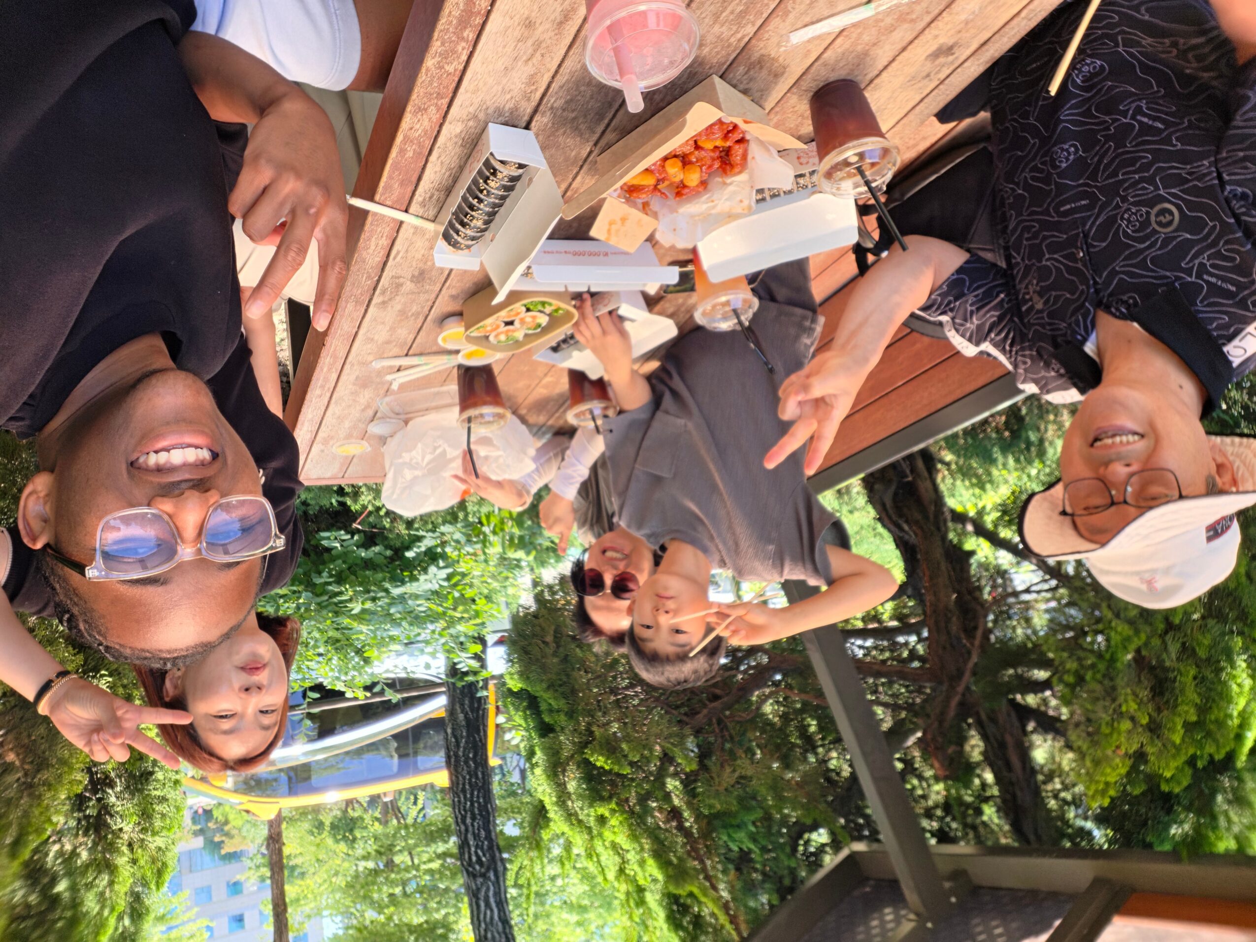 Group of people enjoying food outdoors at a wooden table, smiling and talking, with trees and sunlight in the background.
