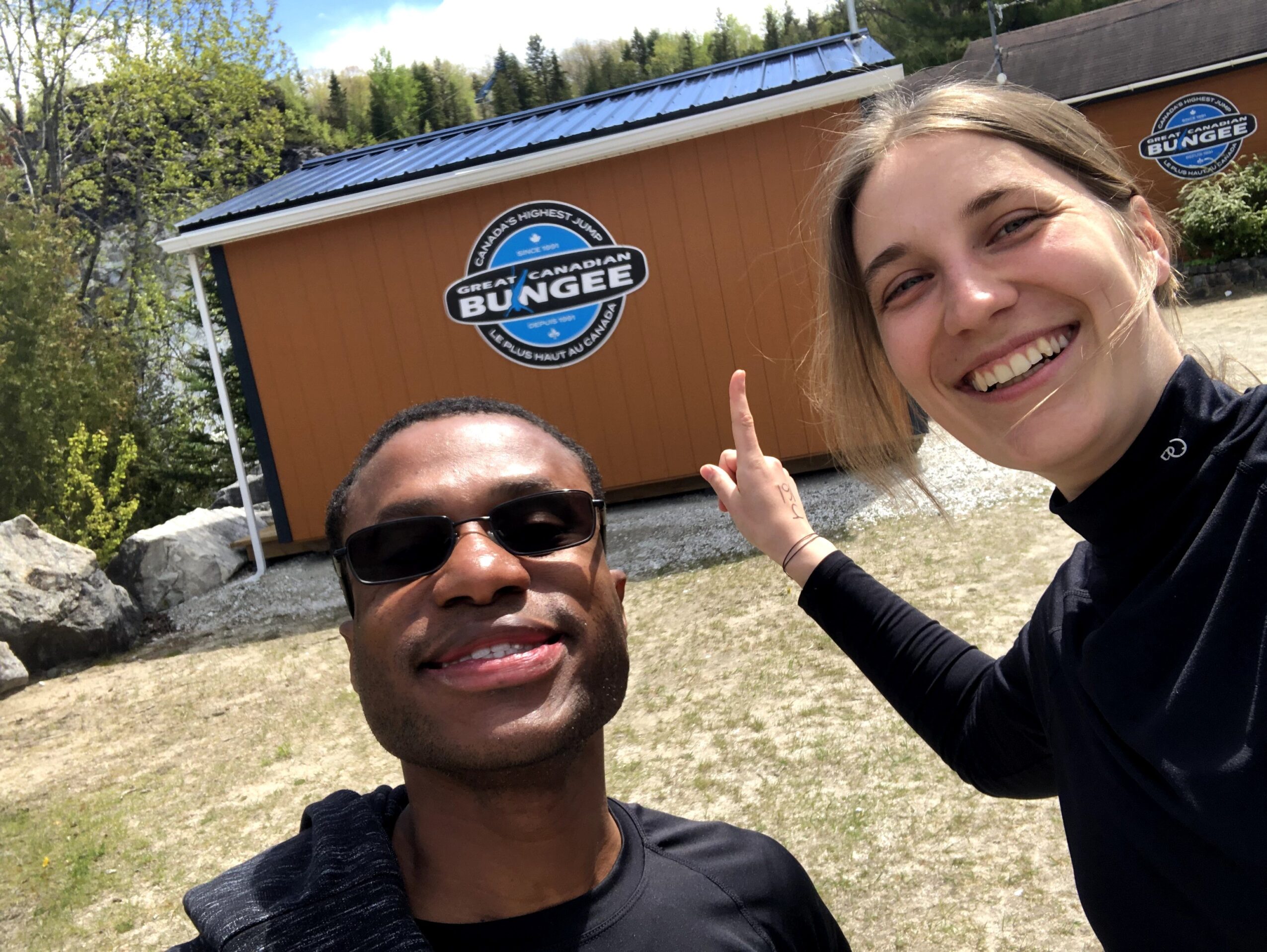Two smiling people outdoors in front of a building with a Bungee Jump sign, one person pointing at the sign.