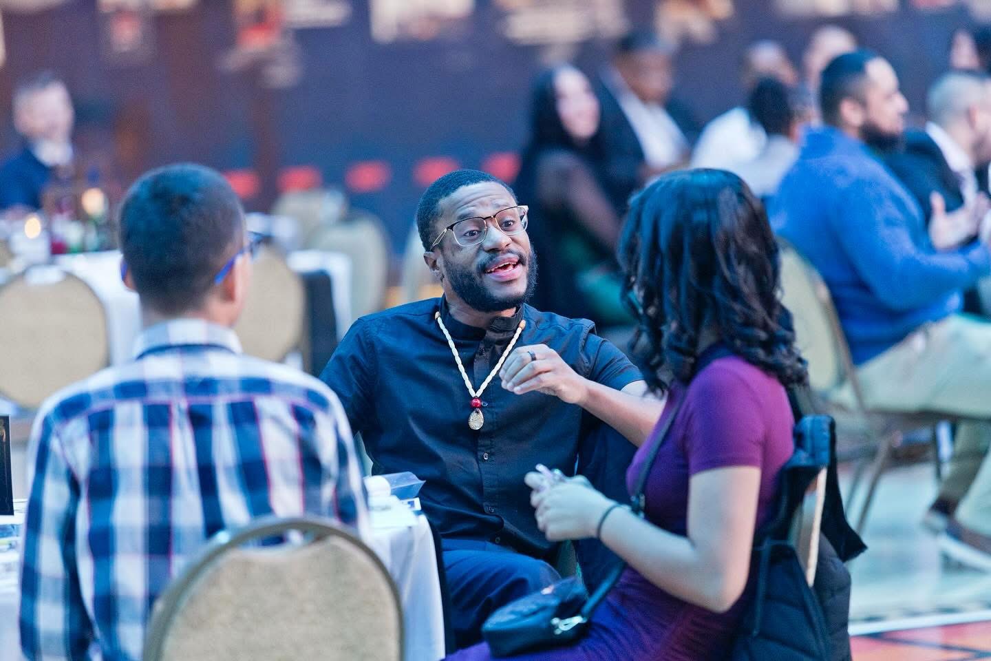 Three people engaged in conversation at a social event, with tables and other guests in the background.