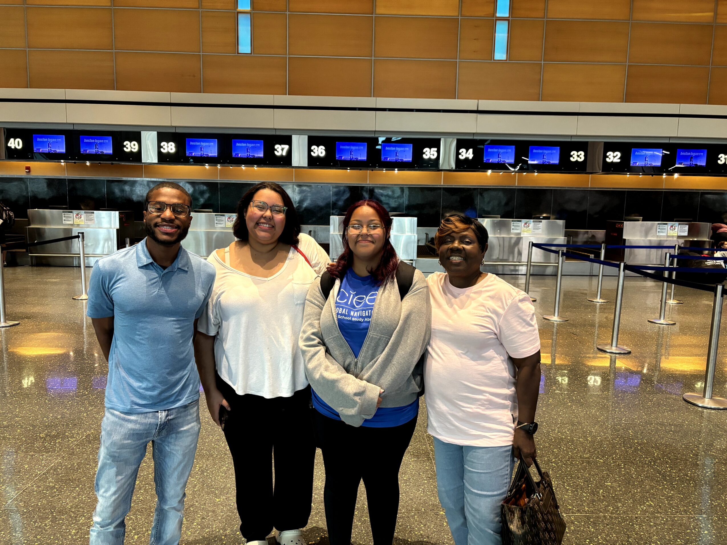 Four people standing in an airport check-in area, smiling, with check-in counters and digital screens in the background.