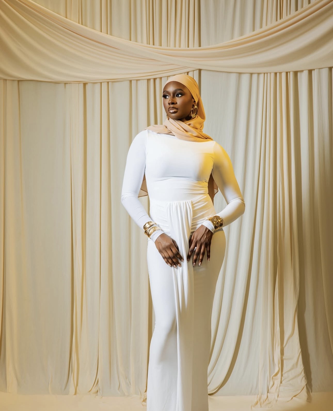 Woman in white dress and headscarf standing in front of cream-colored curtains, looking to the side, with jewelry on her wrists and fingers.