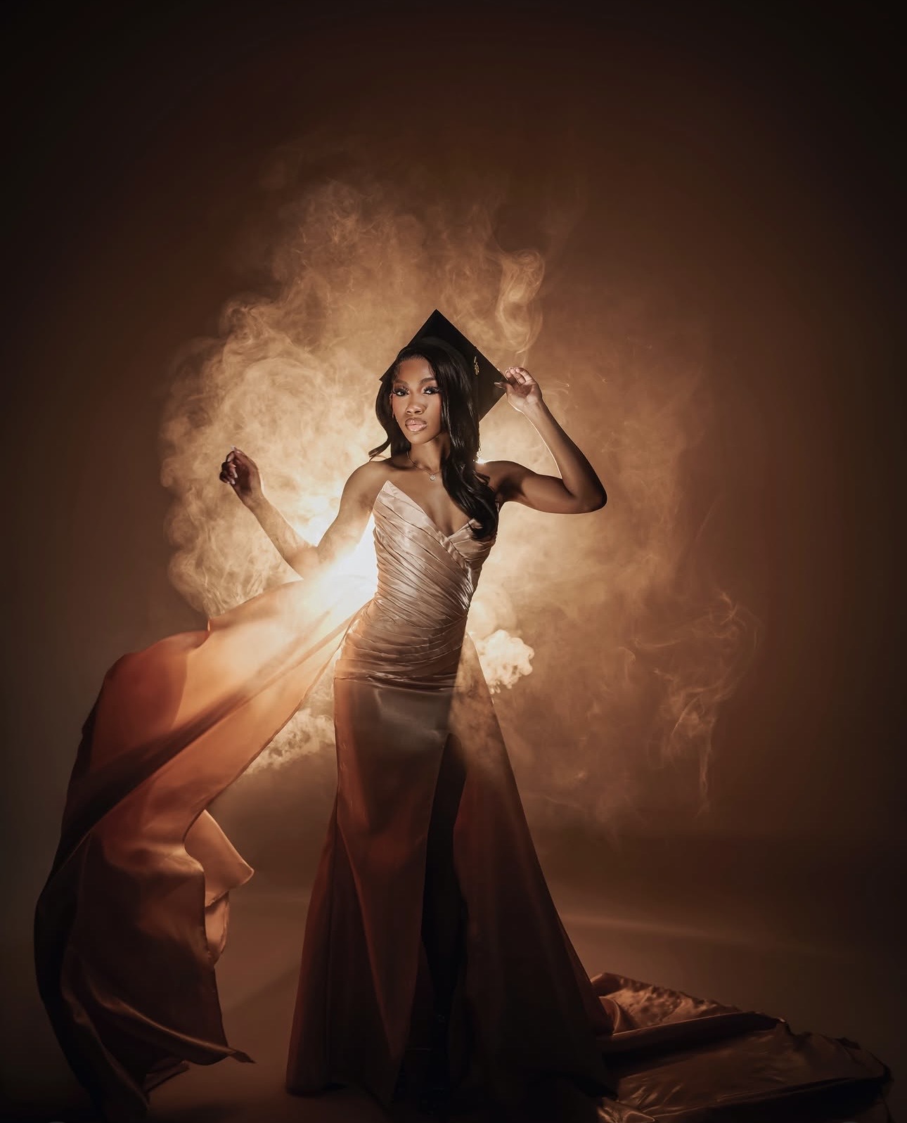 Woman in a gown with a dramatic background of smoke and light, holding a hat, standing on a fabric-covered surface.