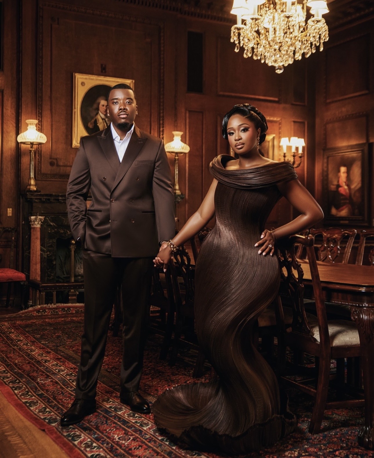 Man in a suit and woman in a black gown holding hands in an elegant room with chandelier and wooden paneling.