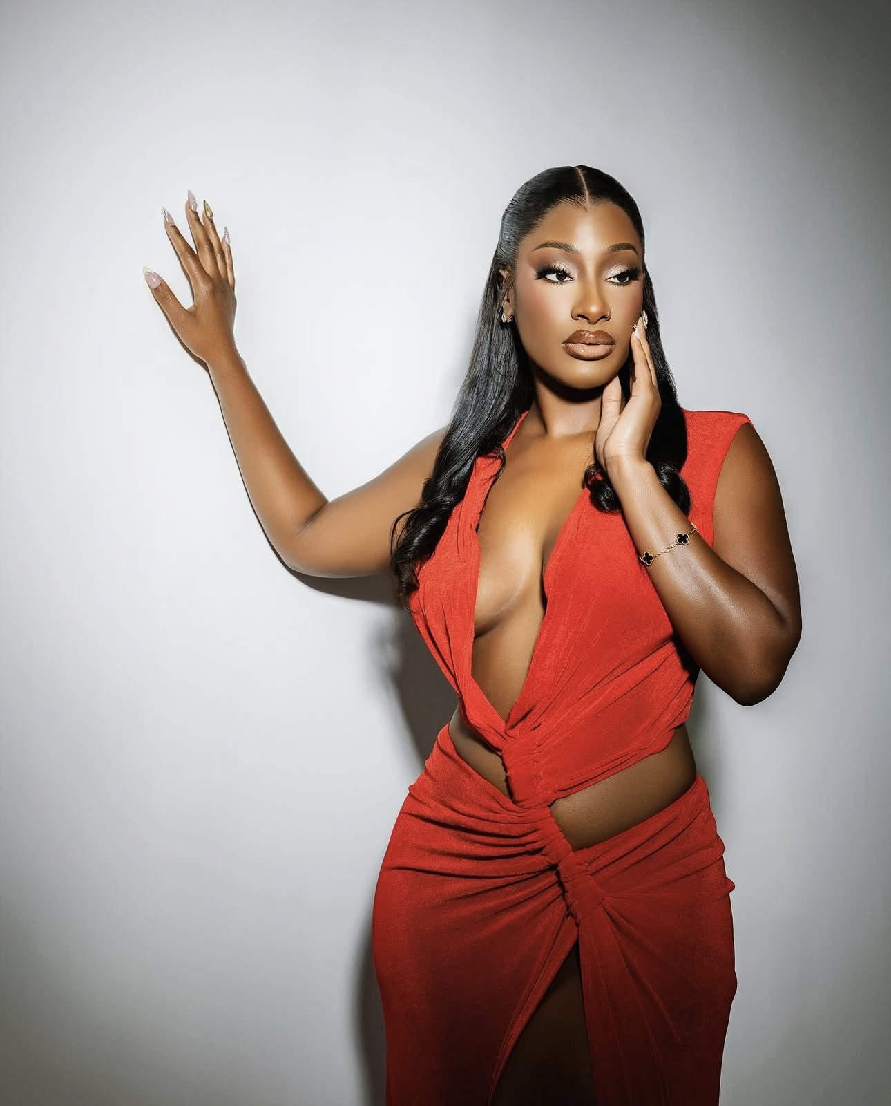 Woman in a red dress with a deep neckline, posing with one hand on her face and the other raised, against a plain background.