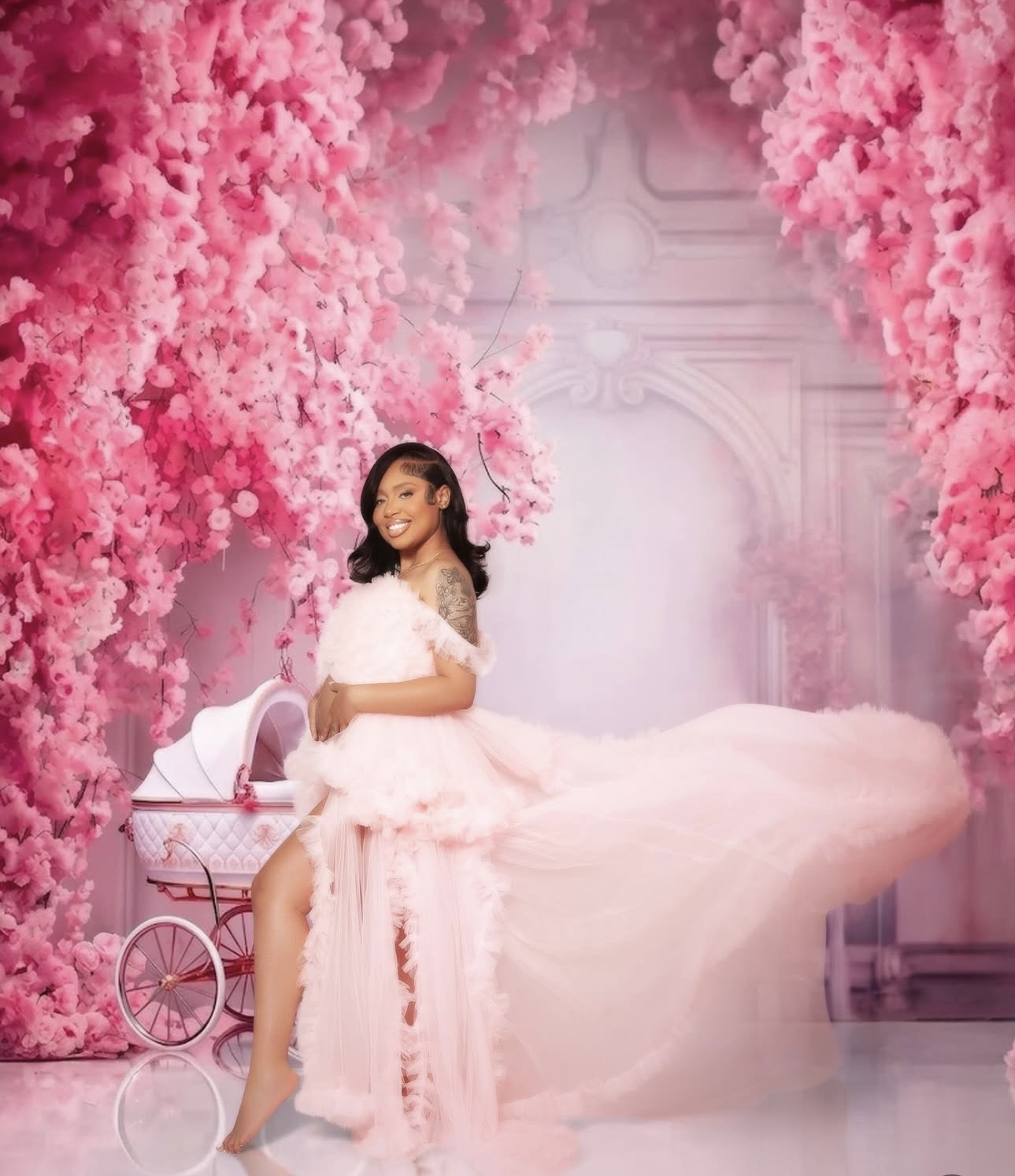 Woman in a pink gown standing next to a pink stroller surrounded by pink flowers and a decorative background.