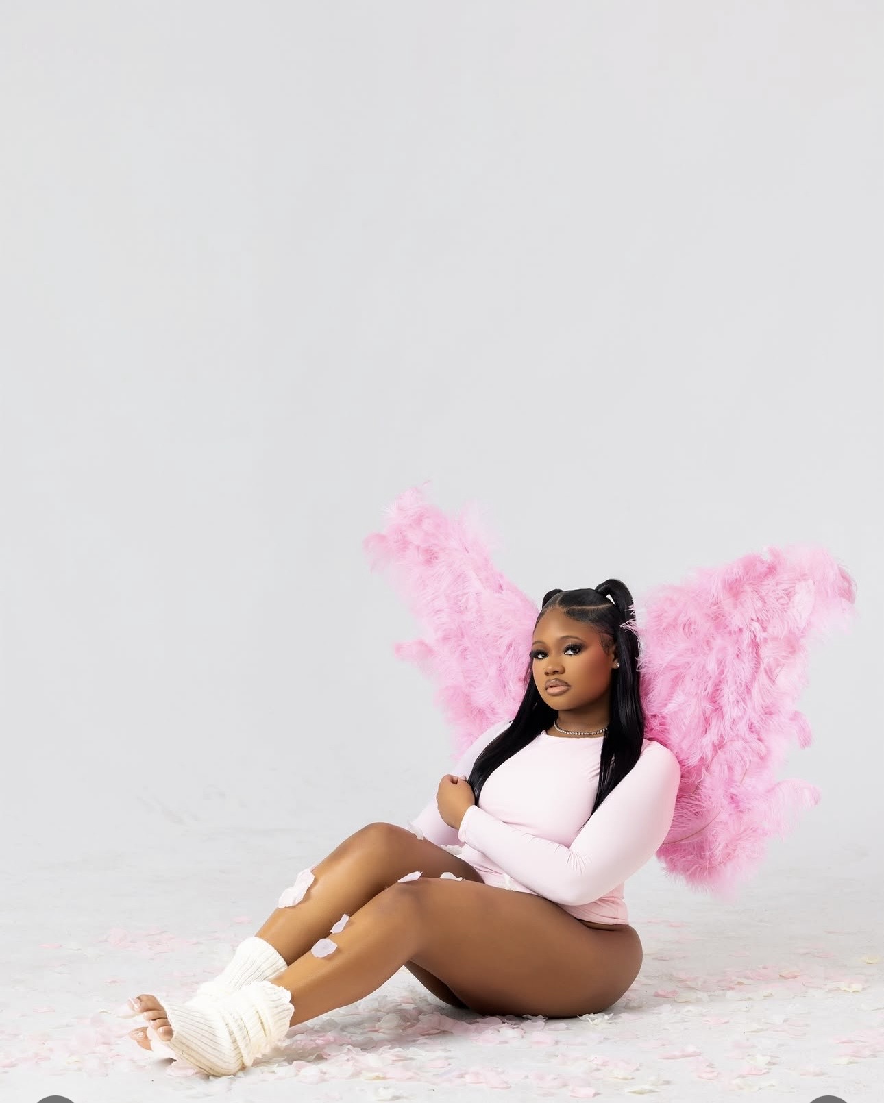 Young woman with pink fluffy wings sitting on the floor, wearing a white top and brown shorts, against a plain background.