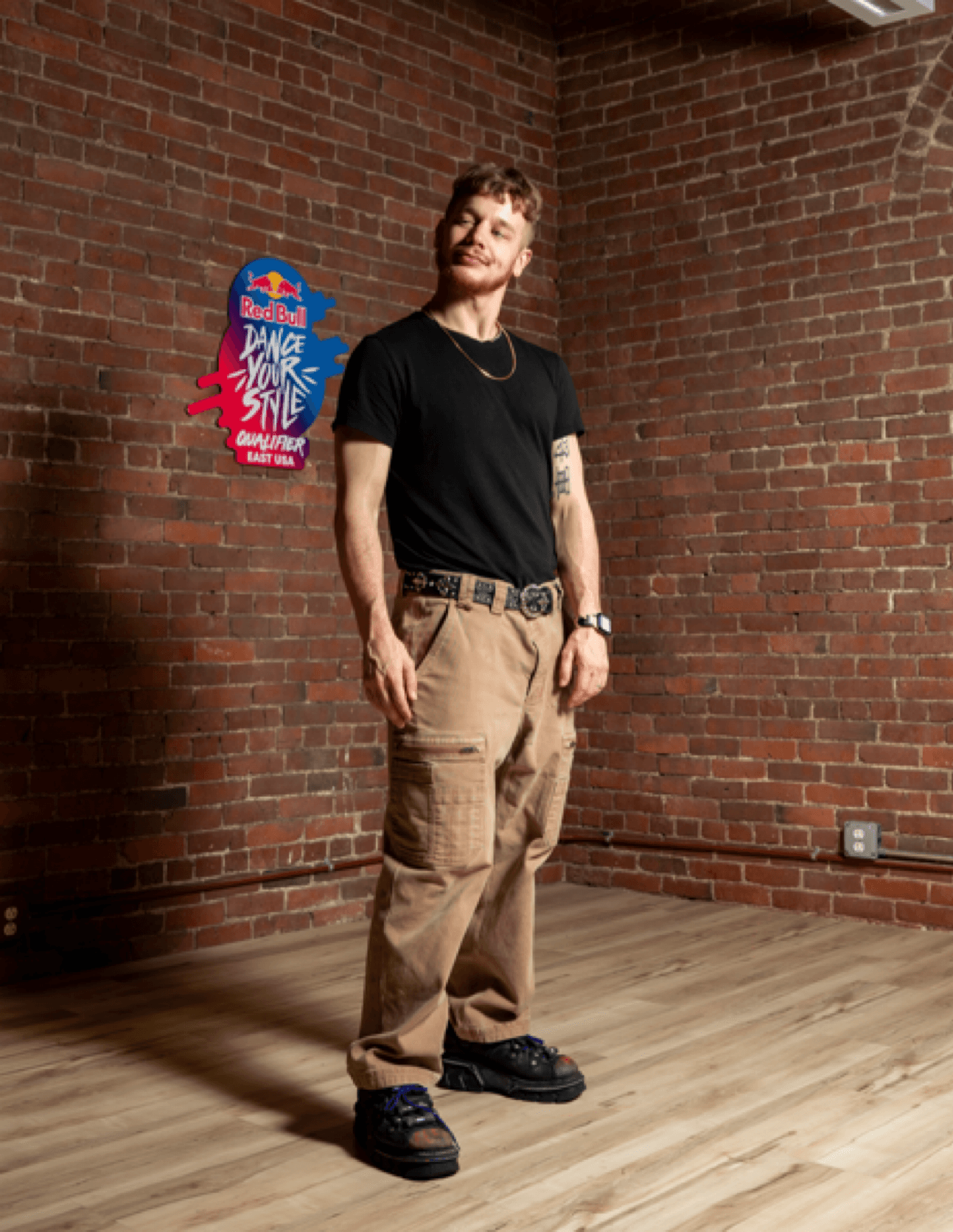 Young man with tattoos, black t-shirt, beige cargo pants, and black shoes standing in front of a brick wall with a colorful poster.