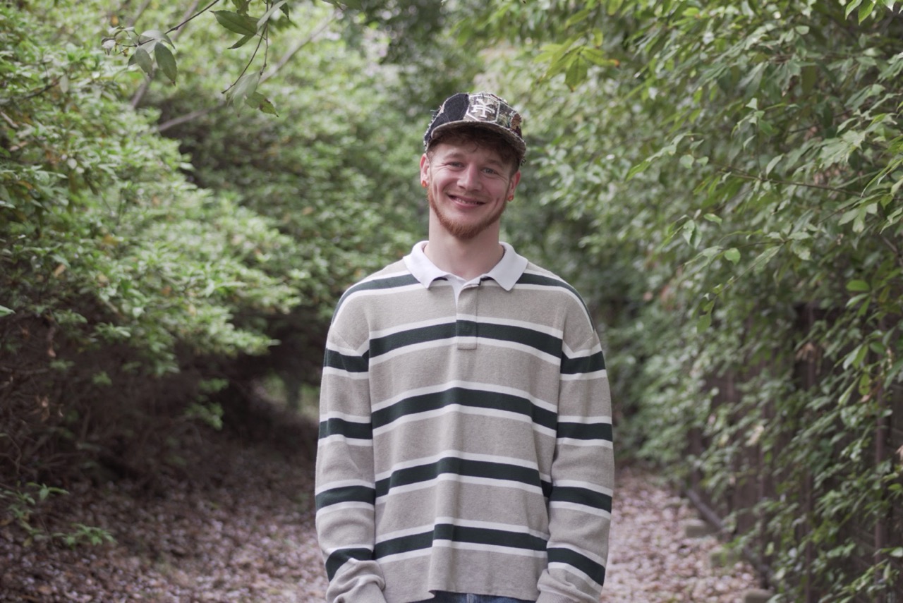 Young man standing outdoors on a dirt path surrounded by green trees and foliage, smiling, wearing a striped long-sleeve shirt and cap.