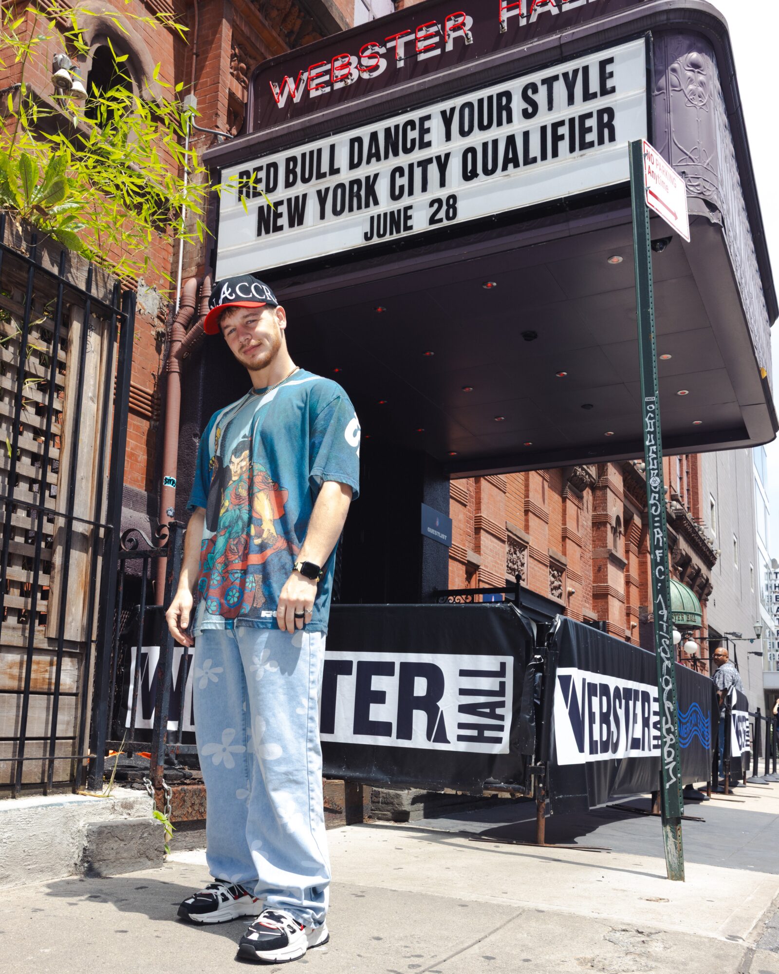 Person standing outside Webster Hall with a marquee displaying event details, wearing a cap, colorful shirt, and jeans.