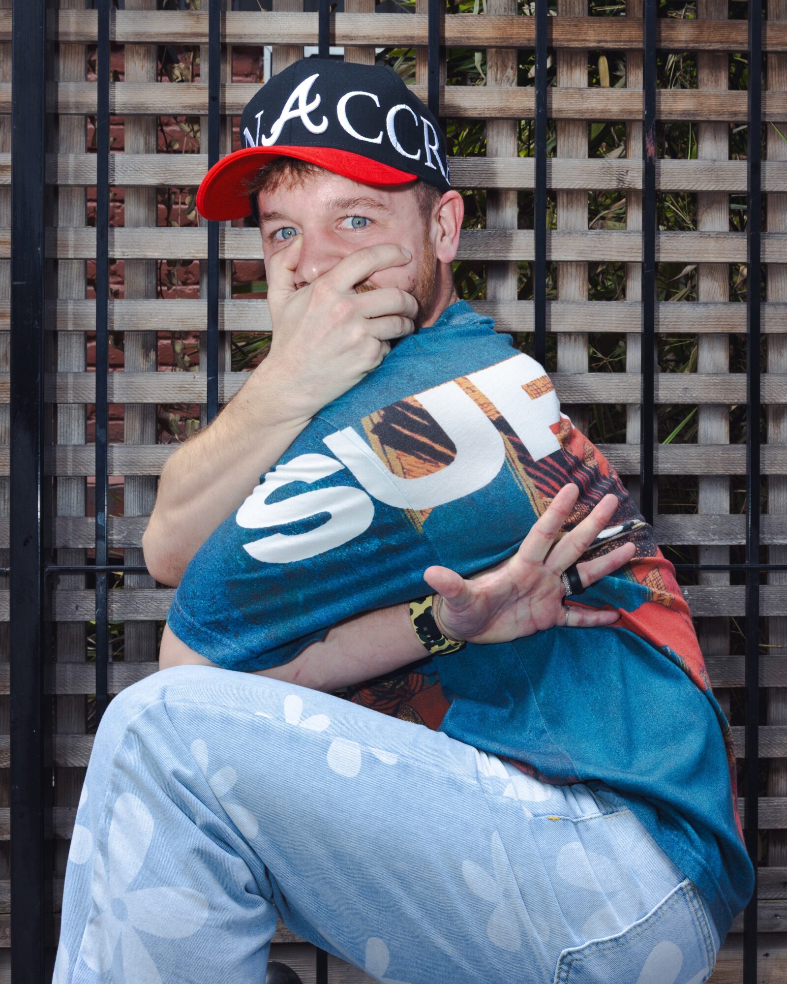 Person sitting outdoors, covering mouth with hand, wearing a cap, colorful shirt, and light-colored pants, in front of a wooden fence.