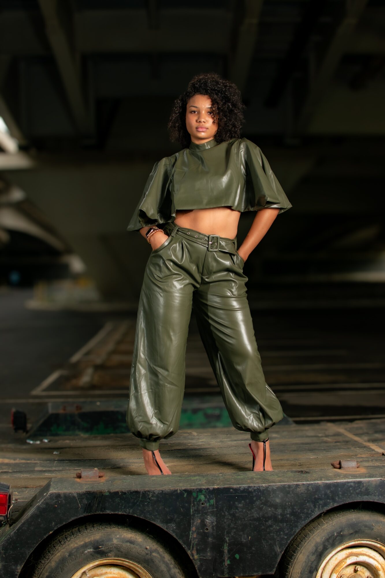 Woman with curly hair in a shiny green crop top and wide-leg pants stands on a platform under a bridge.
