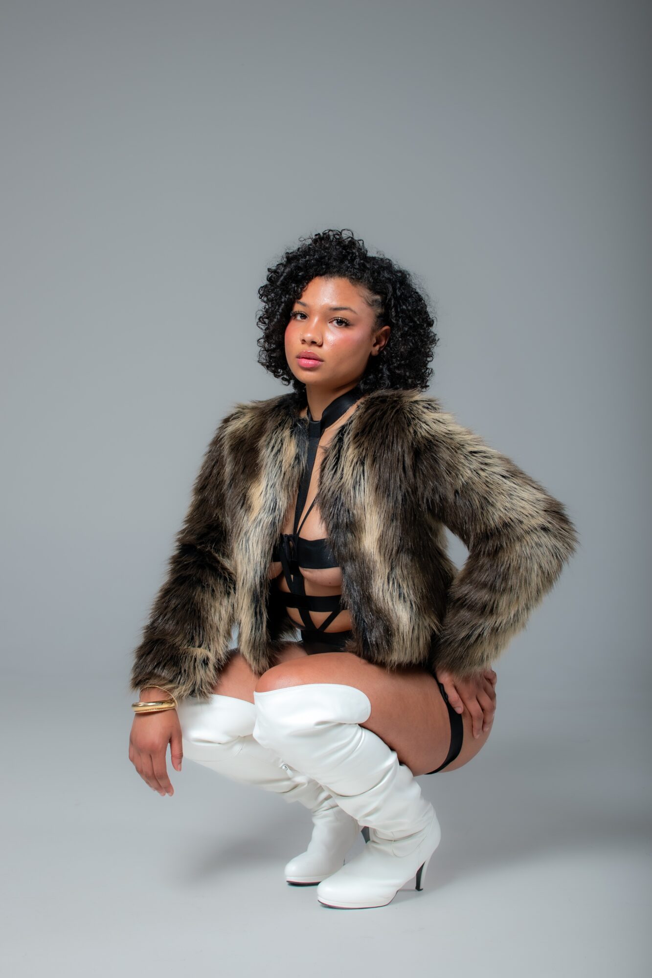 Woman with curly hair wearing a fur coat, black strappy top, white thigh-high boots, squatting against a plain gray background.