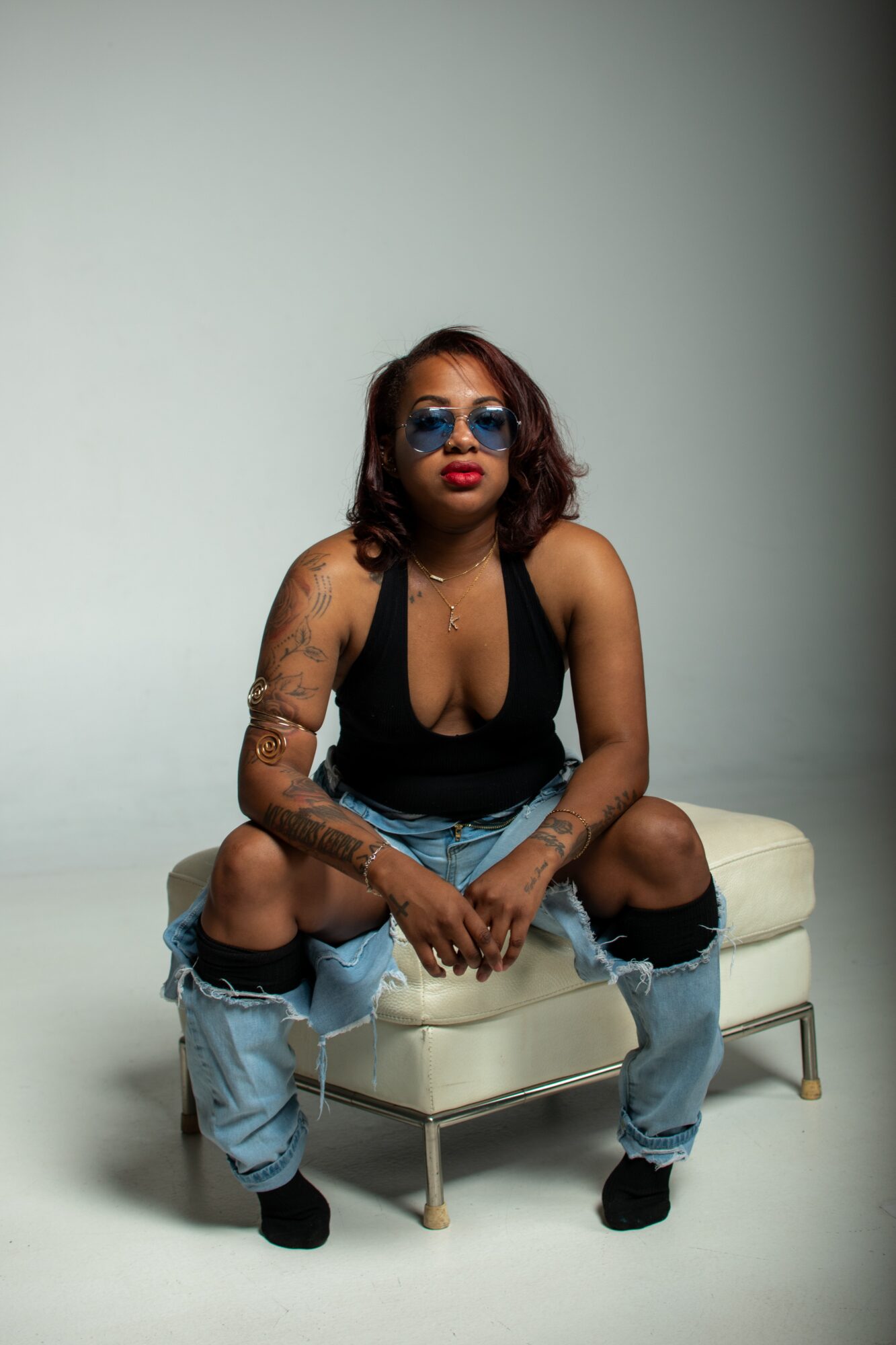 Woman with sunglasses and tattoos sitting on a small white bench, wearing a black top and ripped jeans, against a plain background.