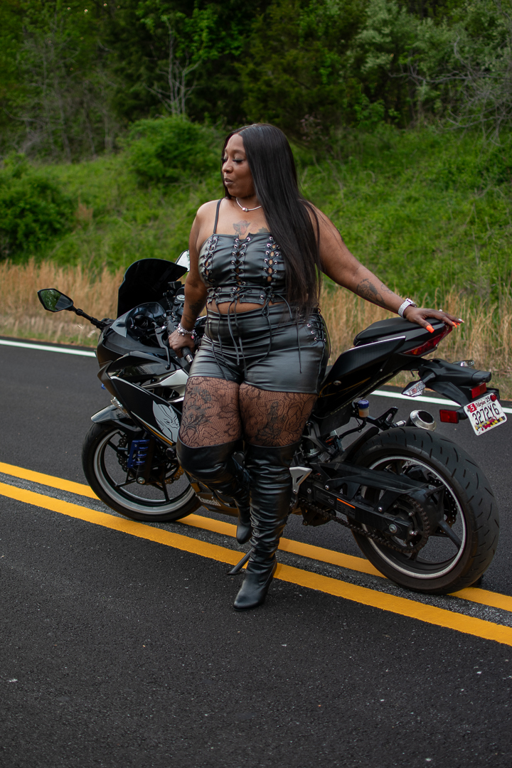 Woman in black outfit standing next to a black motorcycle on a road with green trees in the background.