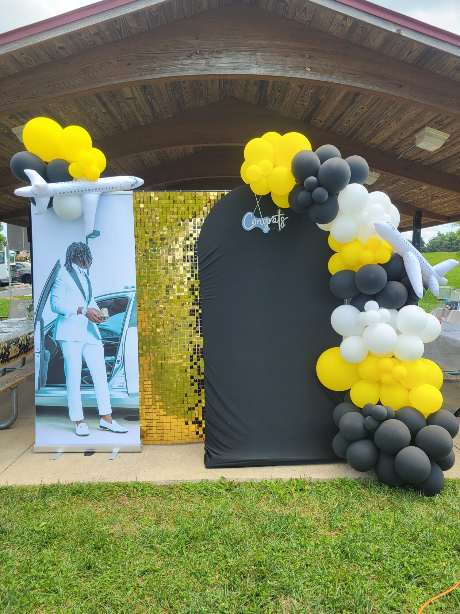 Balloon arch with black, white, and yellow balloons over a black backdrop, and a person in a white costume with a large prop, outdoors.