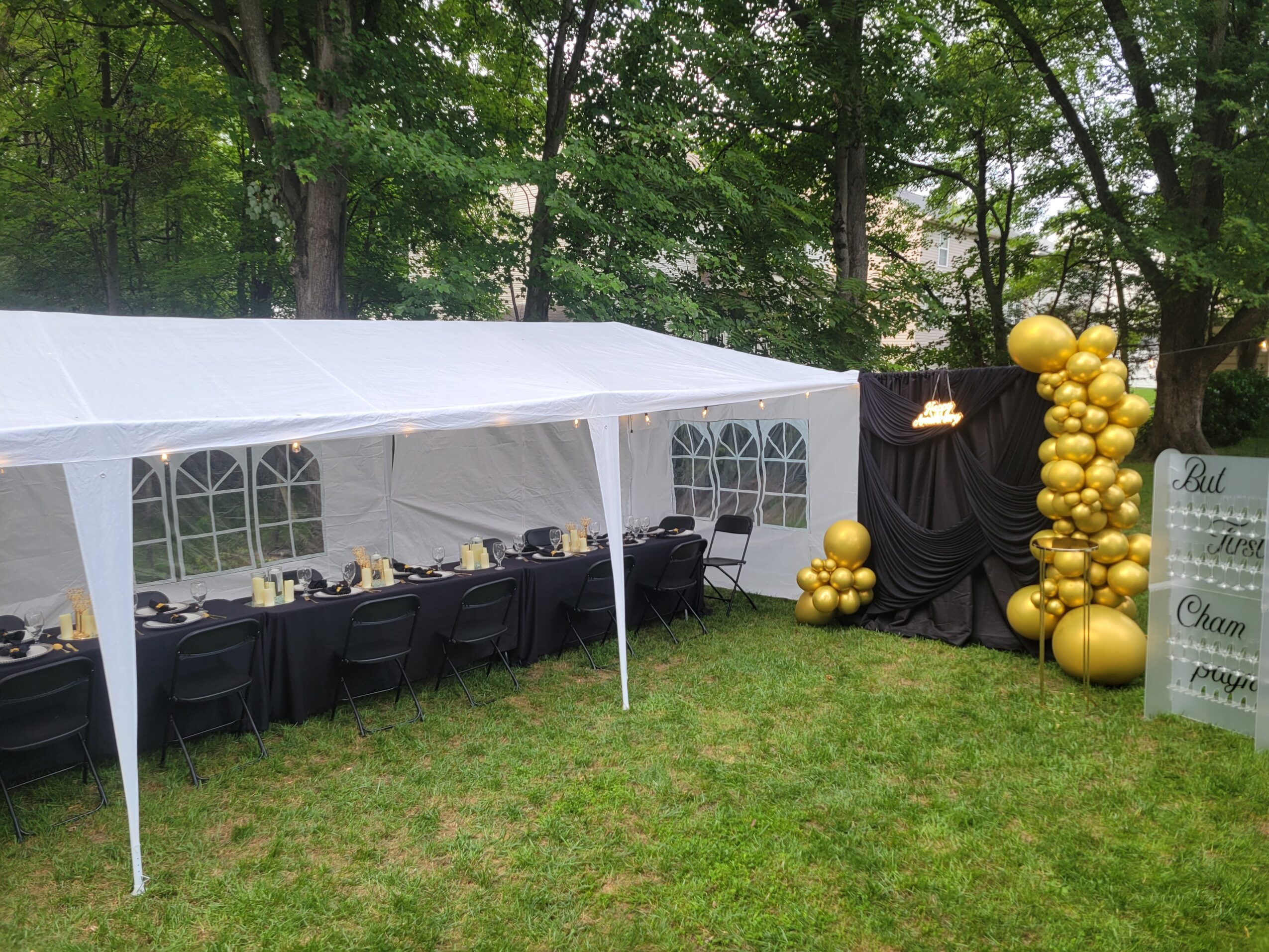 White event tent with black chairs inside, set up outdoors on grass, with gold balloon decorations nearby, surrounded by trees.
