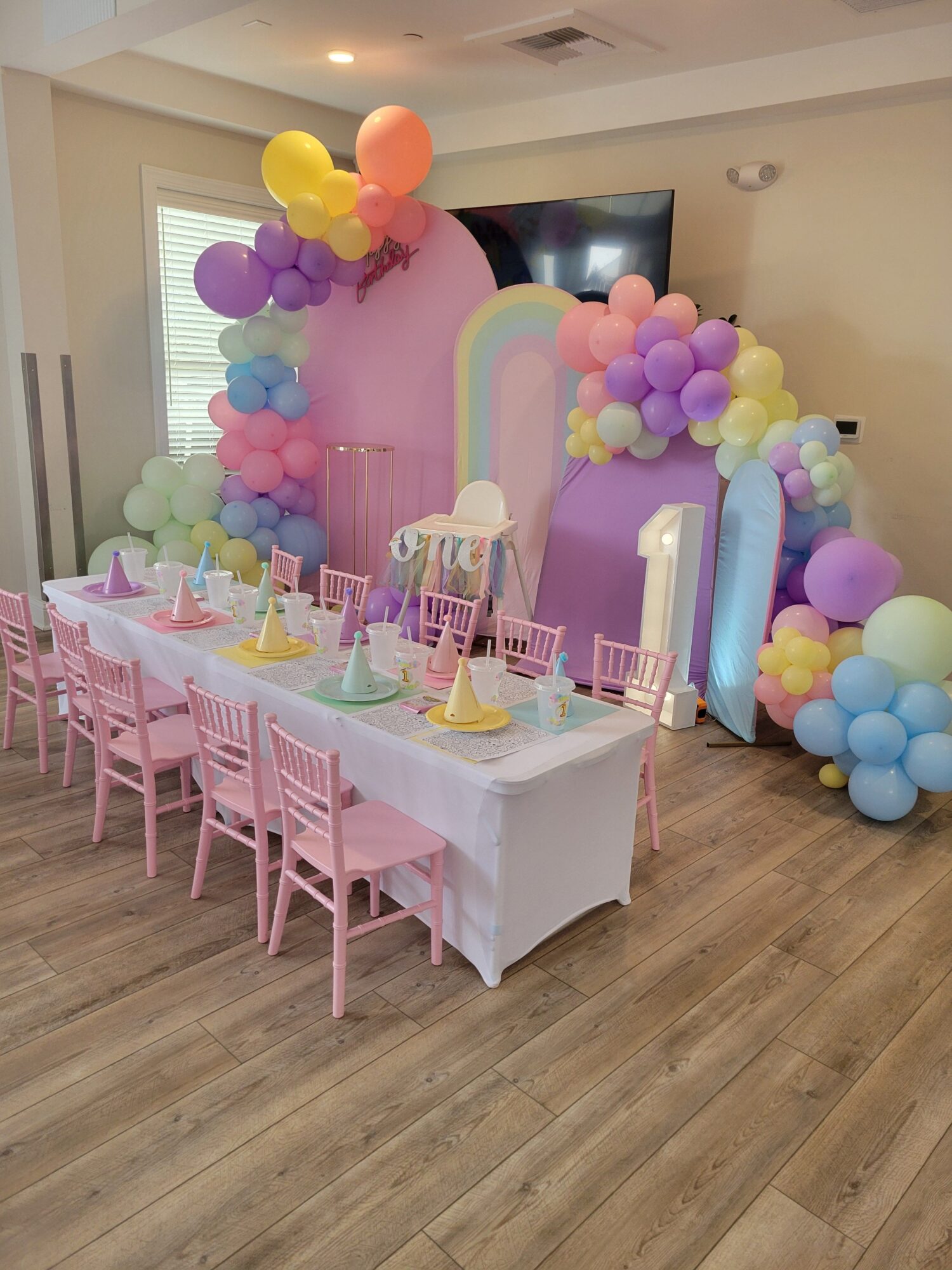 Party table with pink chairs, decorated with colorful balloons and a cake, set in a room with wooden floor.