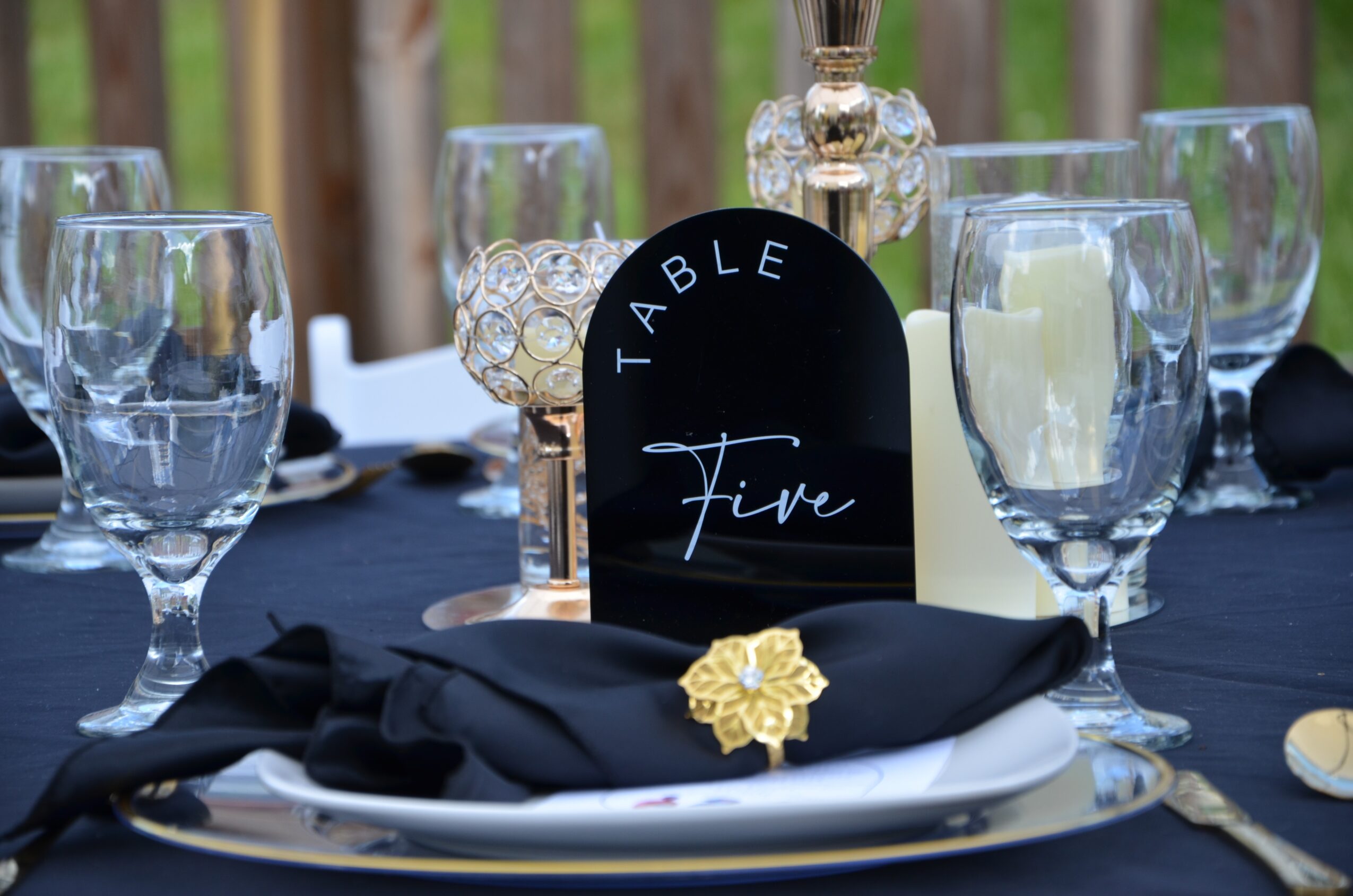 Table setting with wine glasses, a black table number sign, and a black napkin with a gold flower, outdoors.