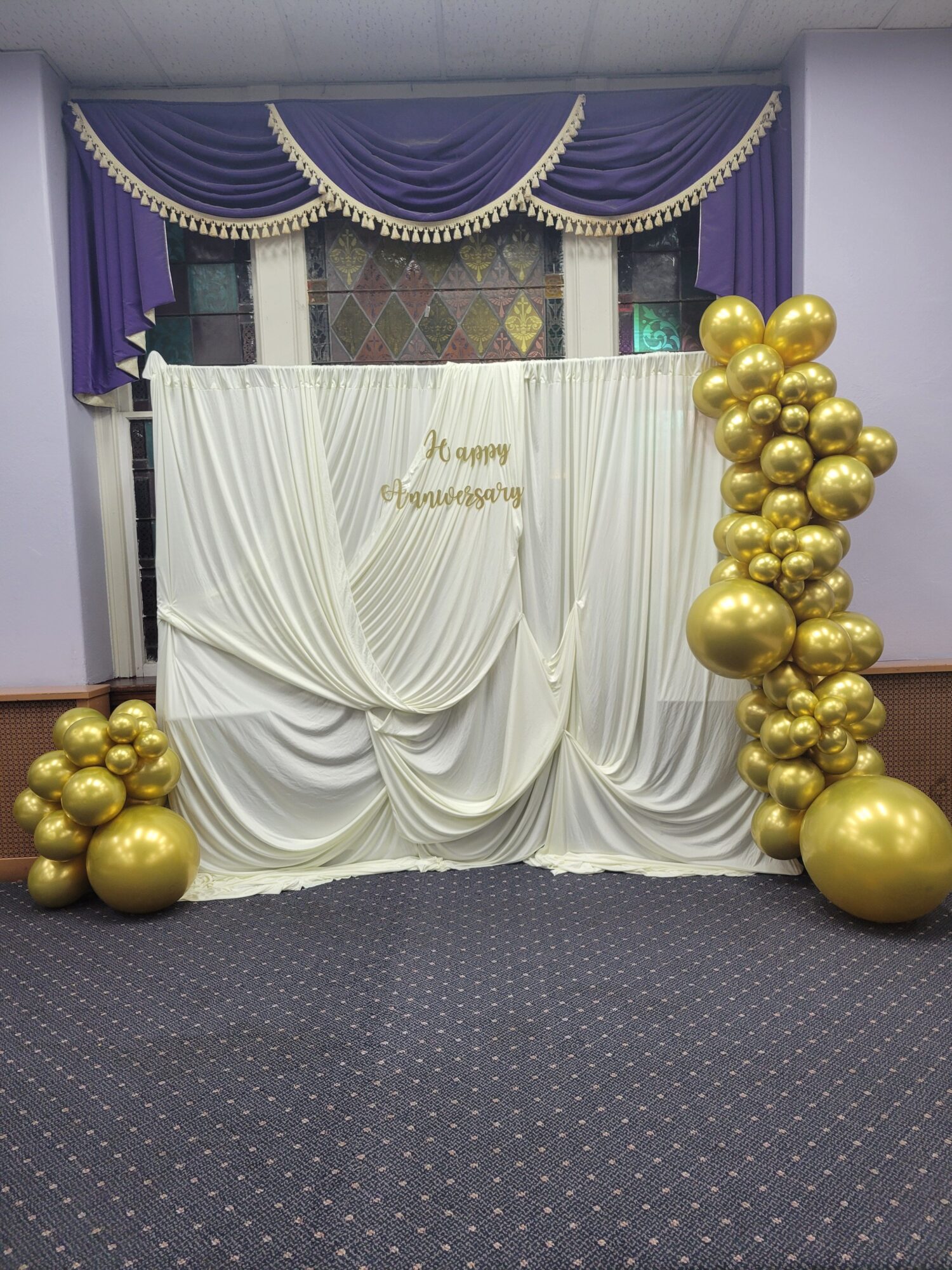 White draped backdrop with gold balloons, purple curtains, and a table with a dark tablecloth, set for celebration.