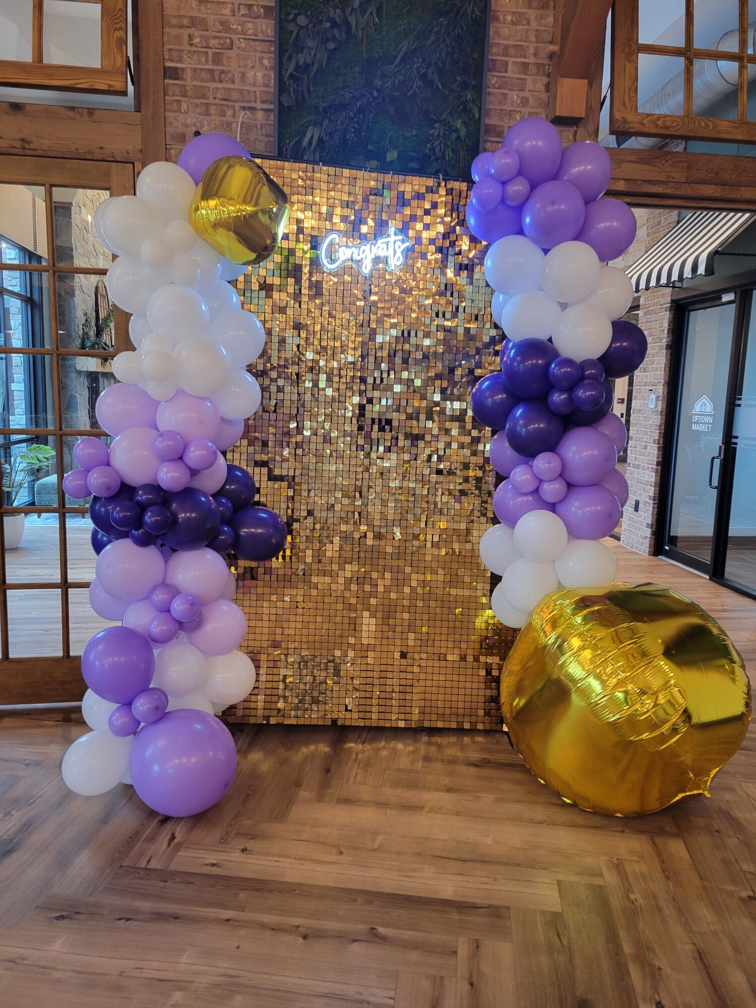 Decorative balloon arrangement with purple, white, and gold balloons, and a gold sequin backdrop, on a wooden floor.