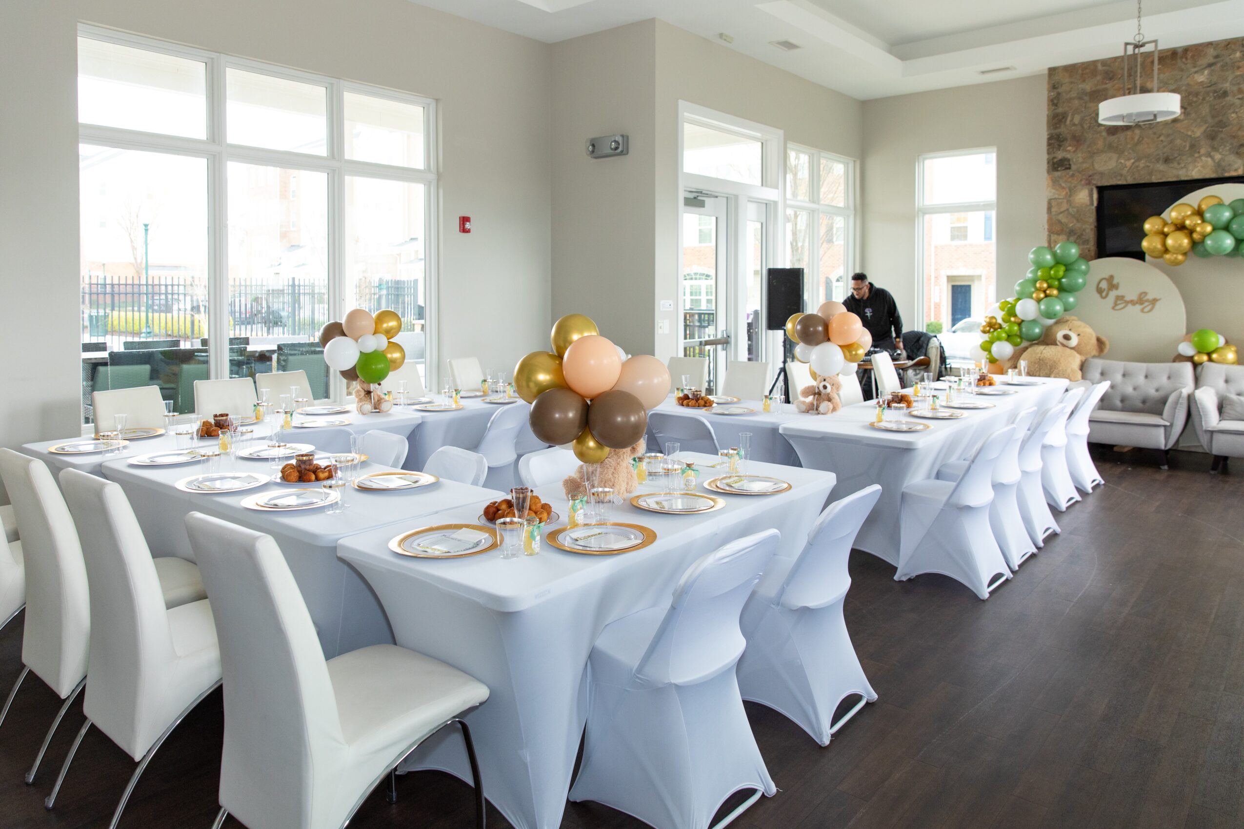 Long banquet table with white tablecloths, decorated with balloons, set for a celebration in a bright, modern room.