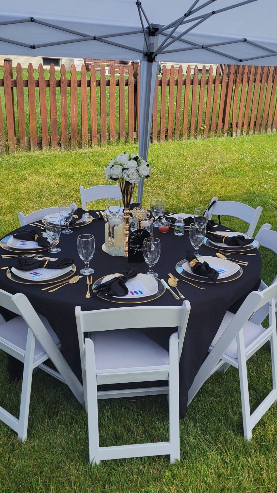 Round table set for outdoor dining with black tablecloth, white chairs, glassware, plates, and a flower centerpiece under a canopy.