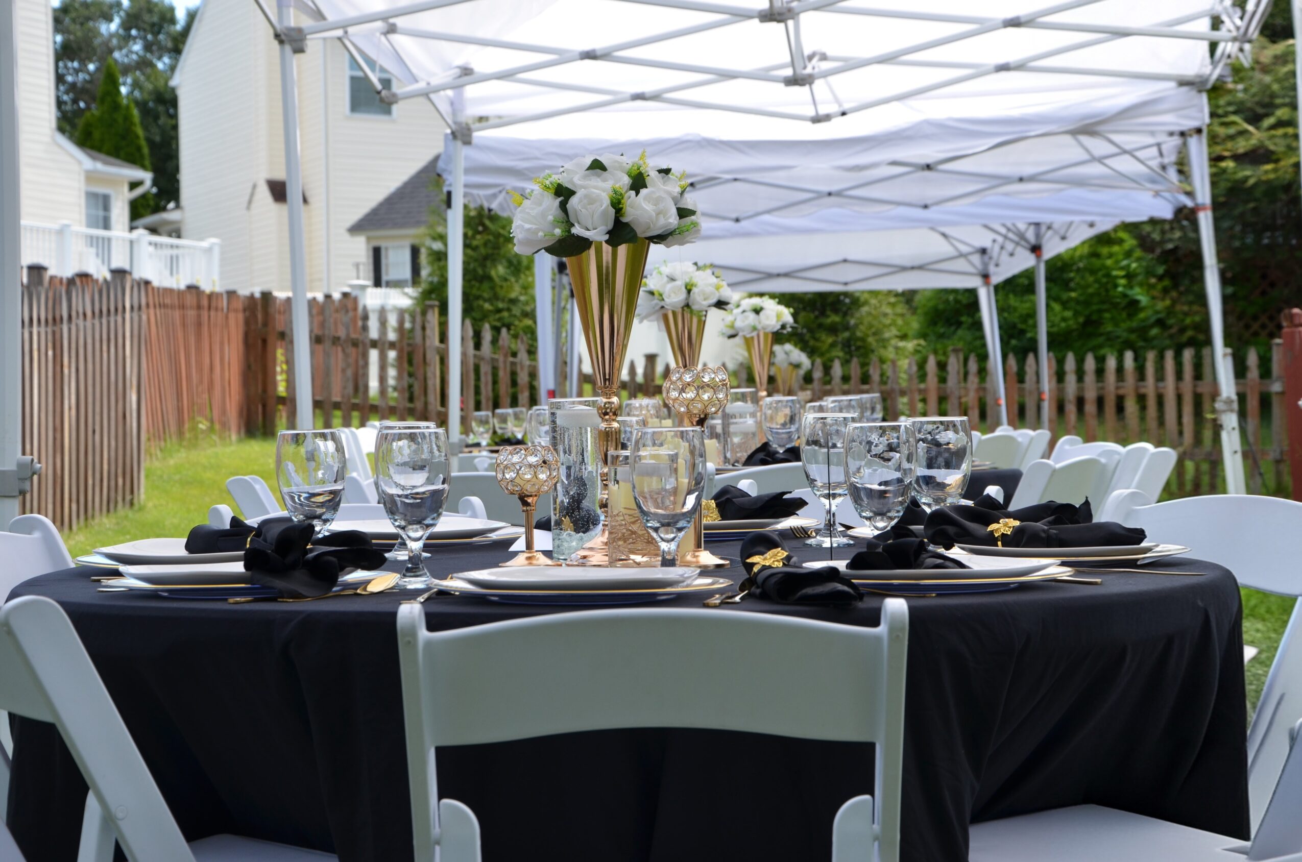 Outdoor table set with black tablecloth, white chairs, floral centerpieces, and tableware under a white canopy.