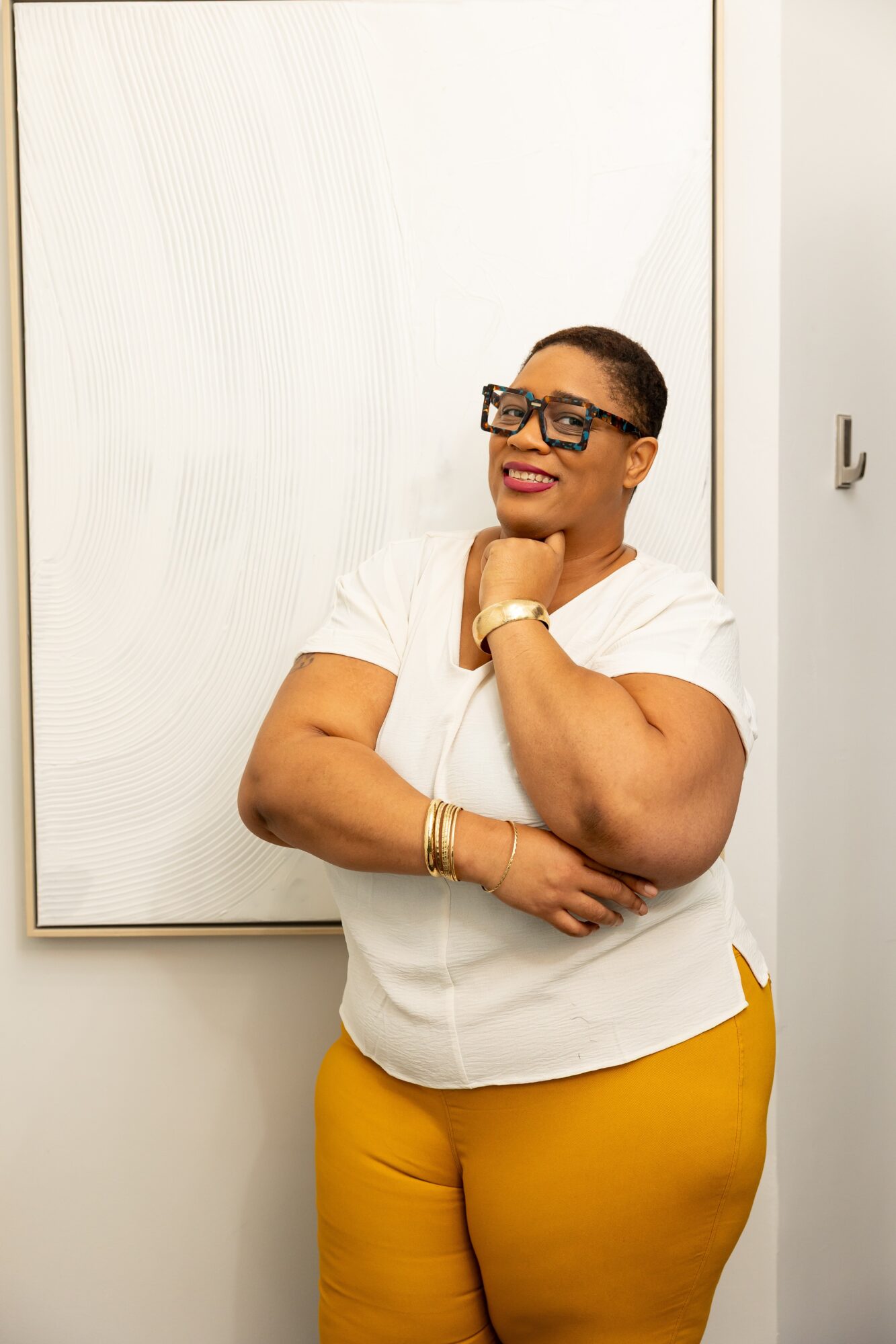 Woman with short hair and glasses, wearing a white top and yellow pants, smiling with arms crossed.