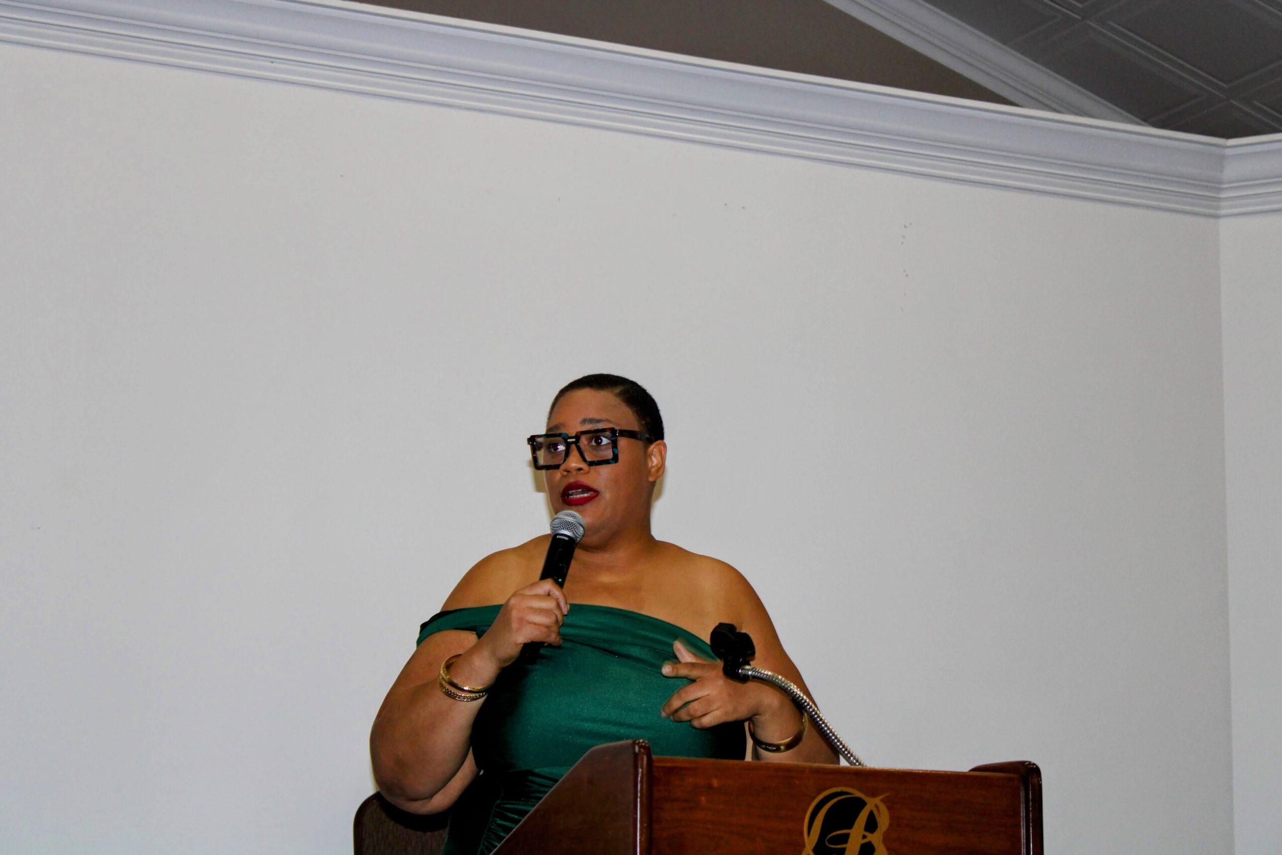 Woman in green dress speaking into a microphone, standing behind a wooden podium, against a plain white wall.