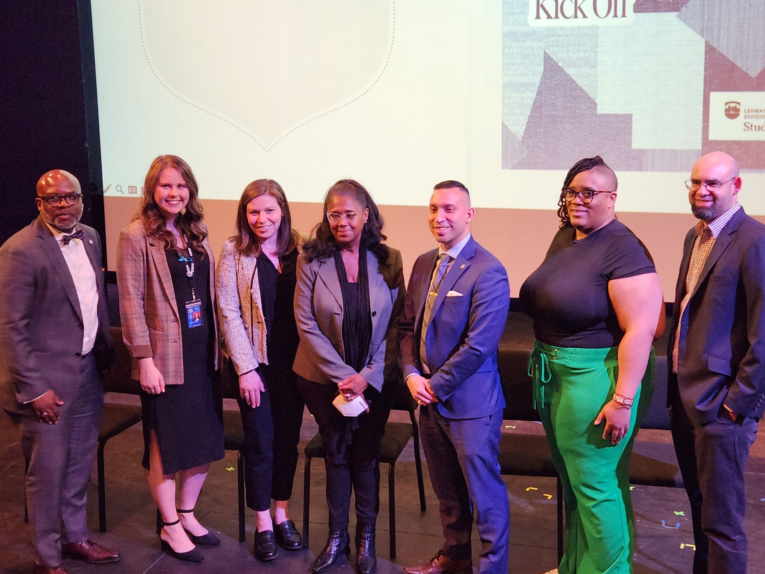 Group of seven diverse people standing on stage, smiling, with a presentation screen behind them.
