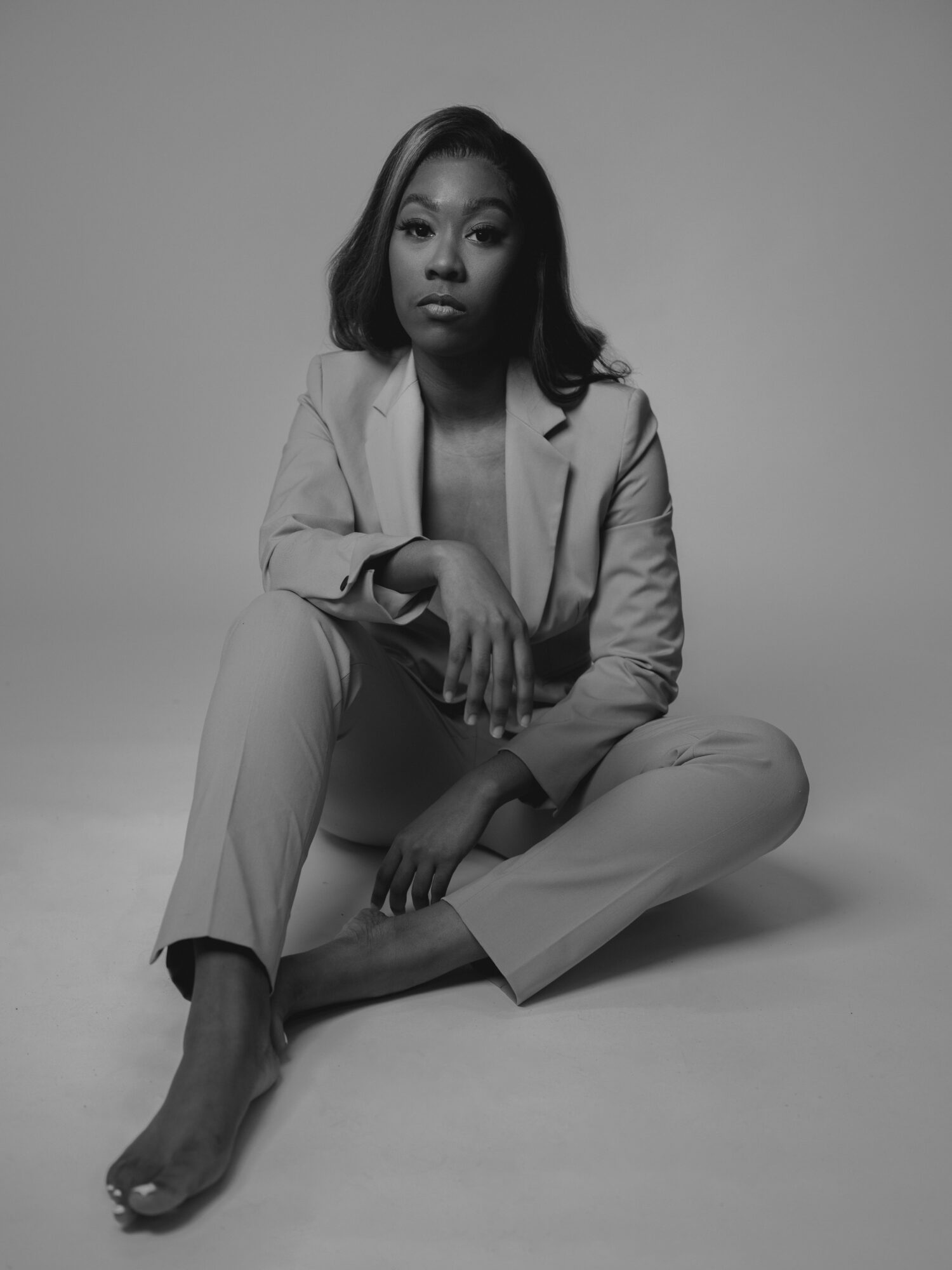 Woman sitting cross-legged, wearing a suit, looking at the camera, in a studio setting, black and white photo.