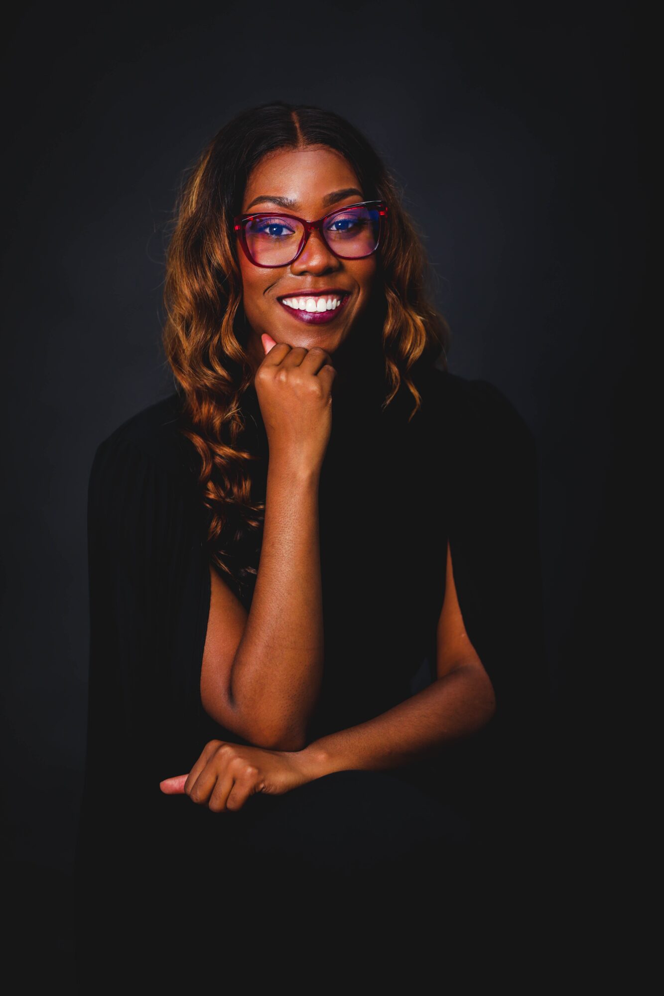 Woman with glasses smiling, resting chin on hand, dark background, curly hair, wearing black top.