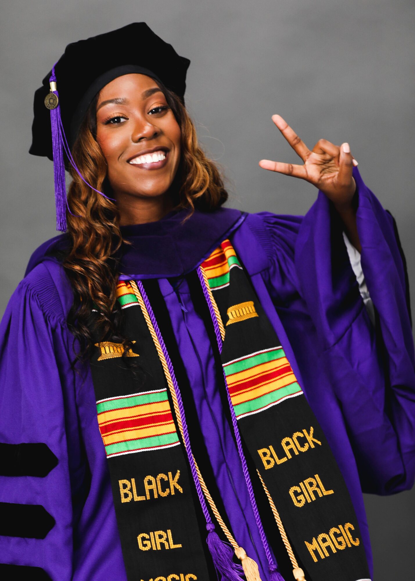 Young woman in graduation gown and cap, smiling, making a peace sign, wearing a colorful stole and sash, gray background.