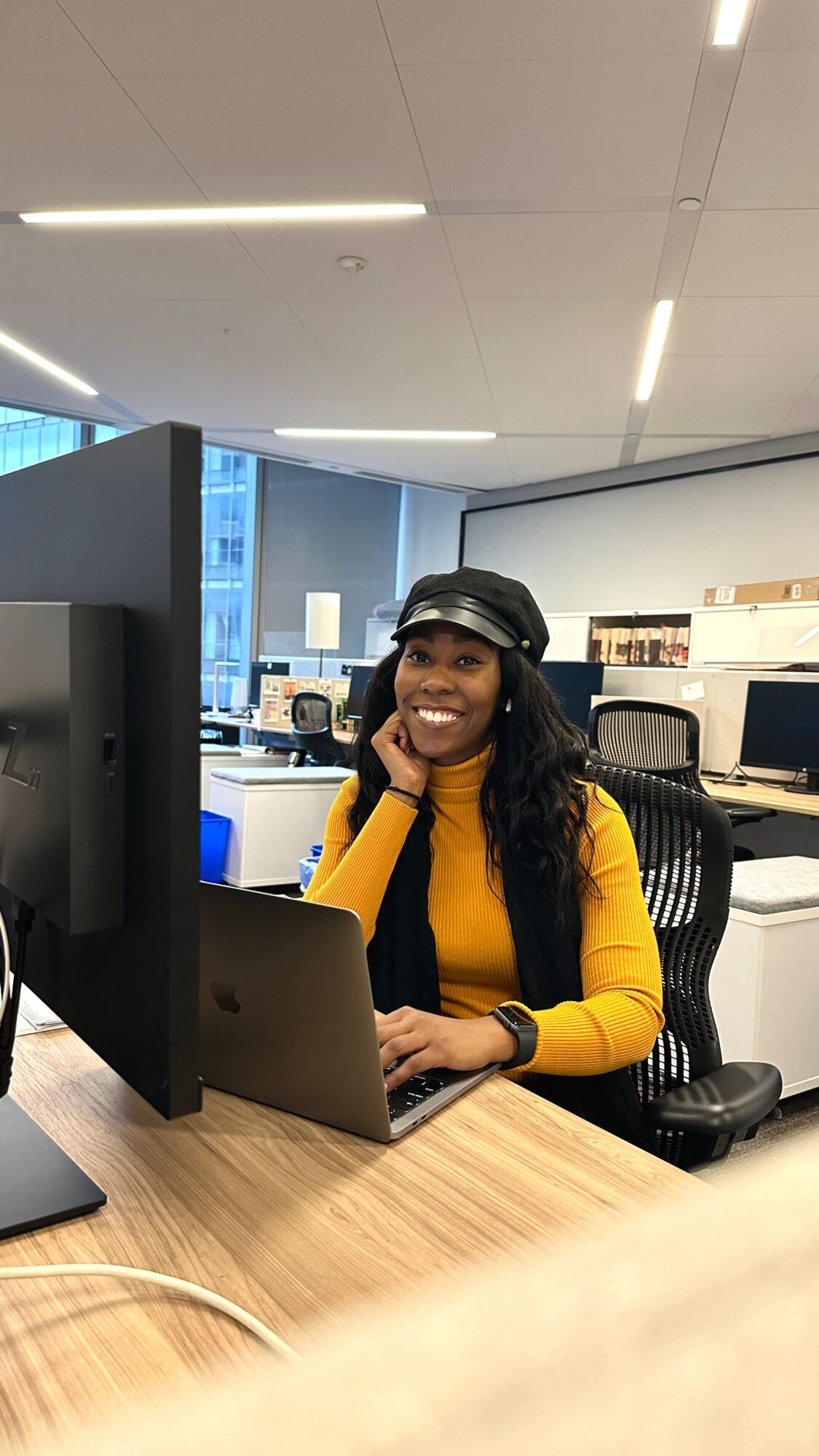 Smiling woman in yellow sweater and black hat working at desk with computer in office.