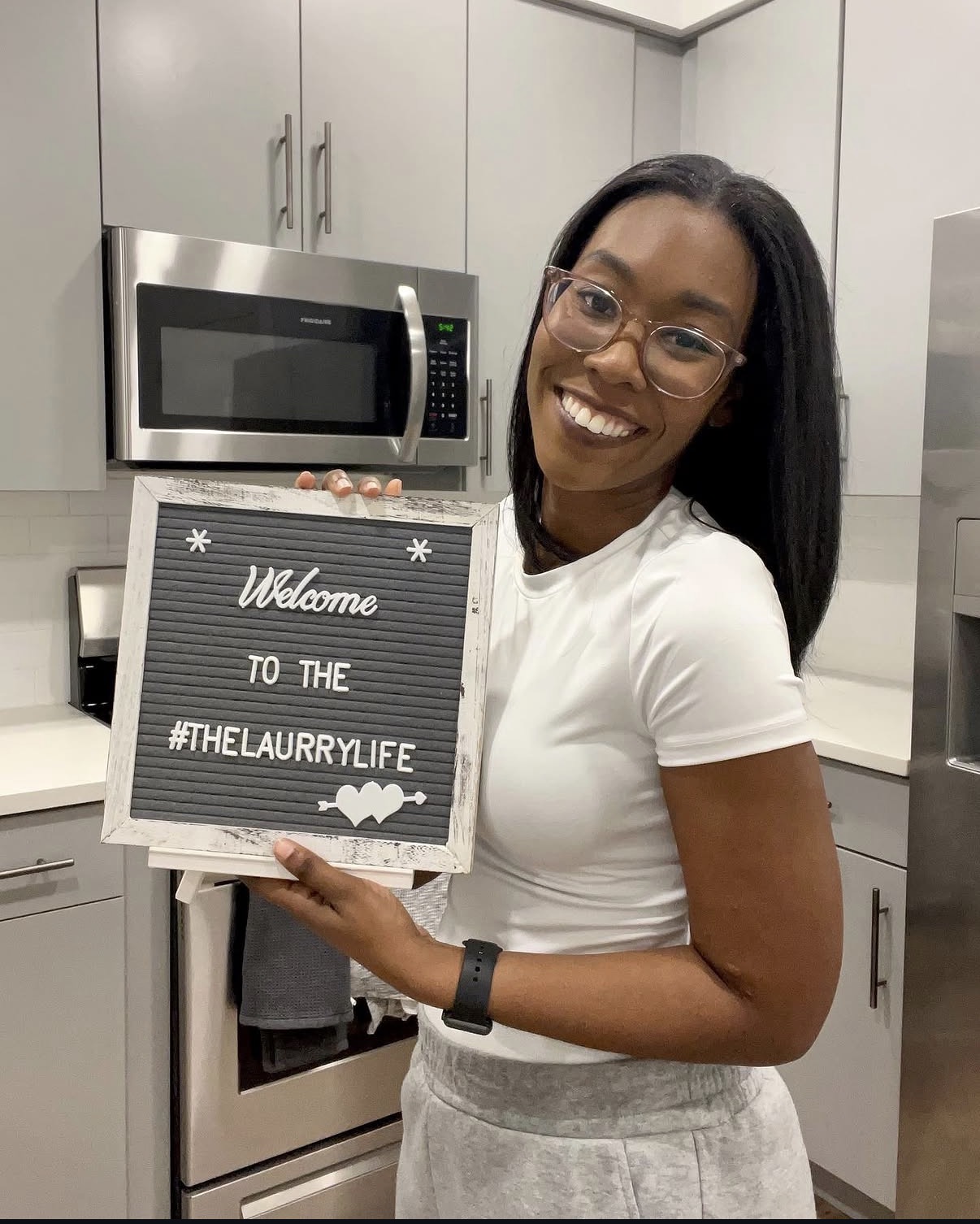 Smiling woman holding a welcome sign in a kitchen with gray cabinets and a microwave.
