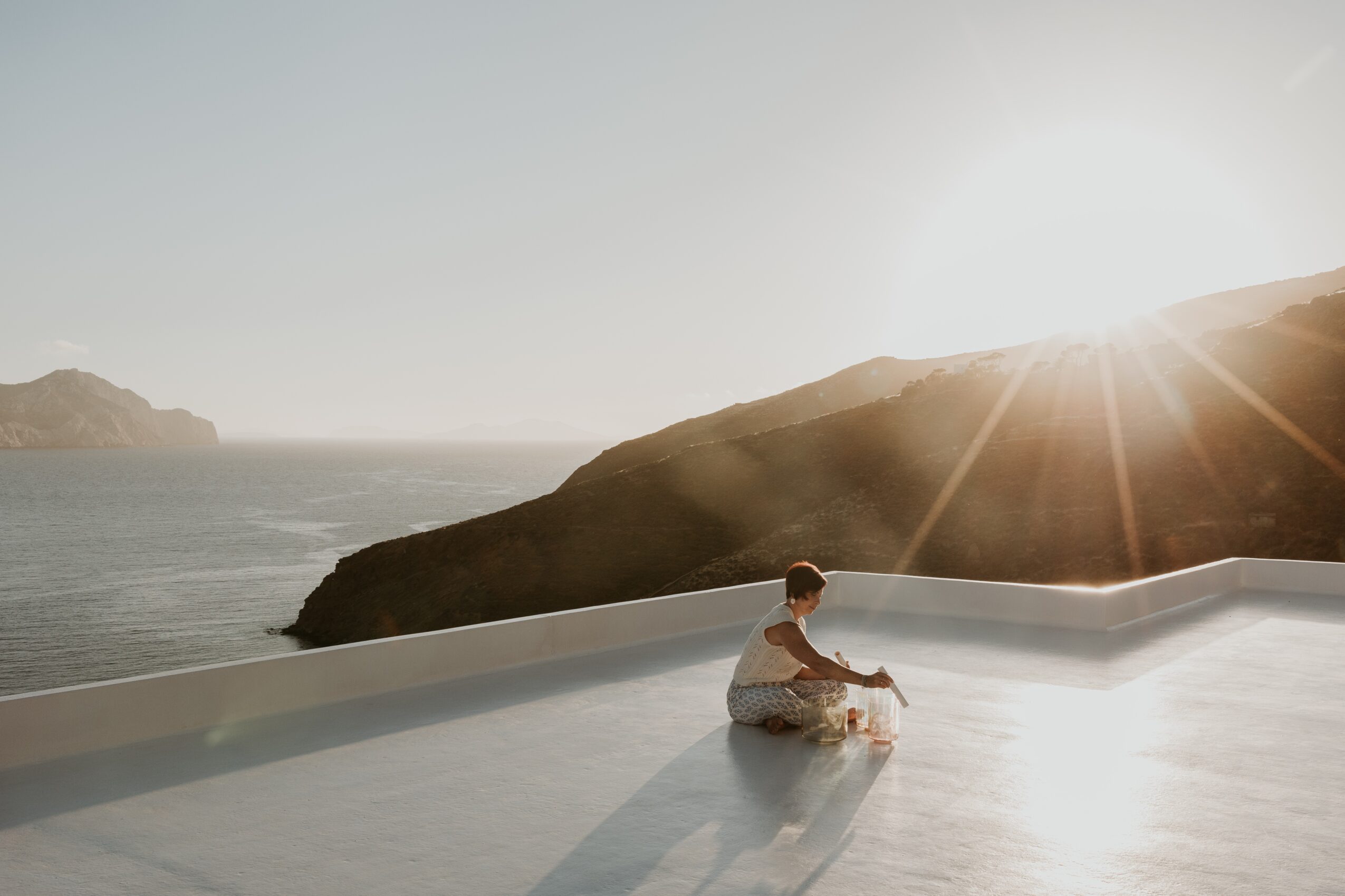 Child sitting on a white surface near water with a mountain in the background and the sun shining brightly.
