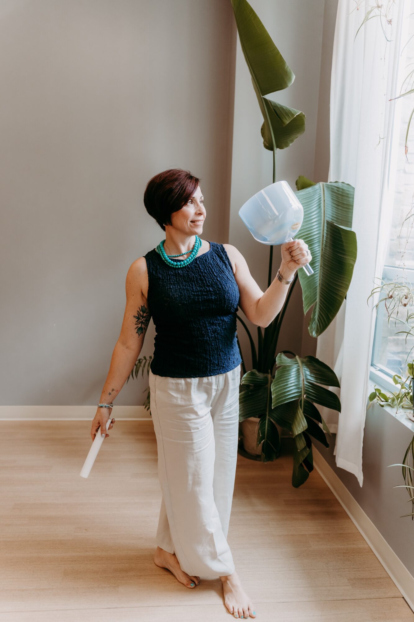 Woman with short hair, wearing a dark sleeveless top and white pants, holding a white hat near a large plant by a window.