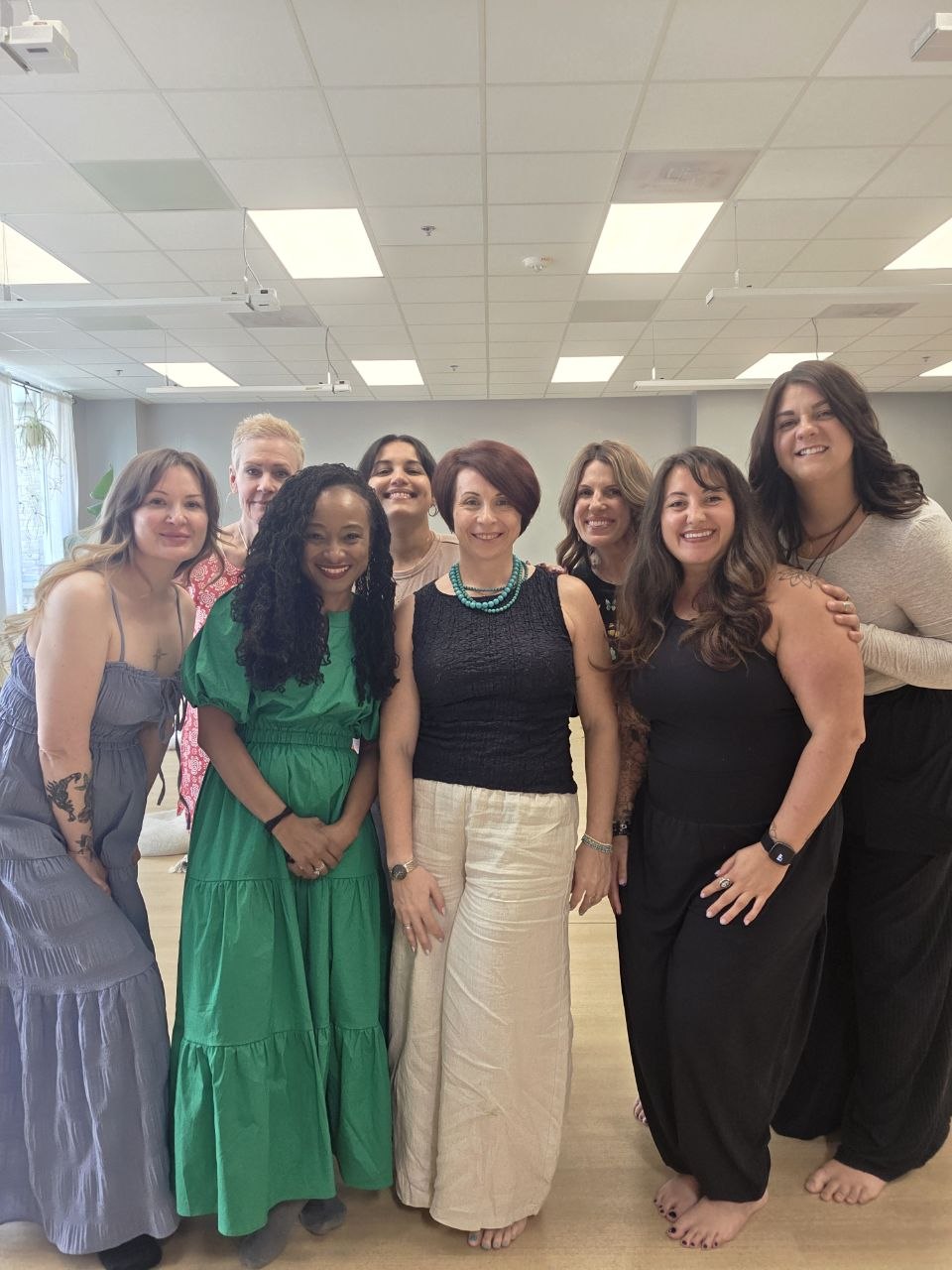 Group of eight women standing together indoors, smiling, with ceiling lights above, some wearing dresses and casual clothing.