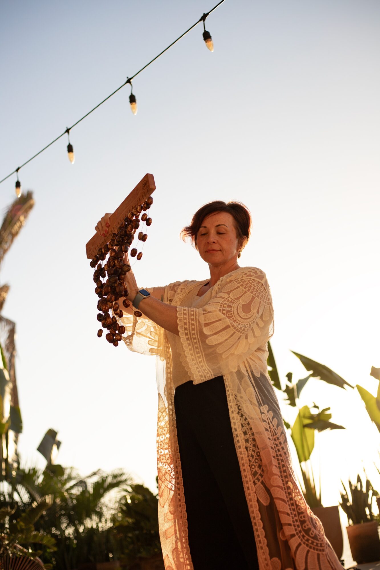 Woman holding a string of dried flowers outdoors during sunset, with string lights overhead and plants in background.