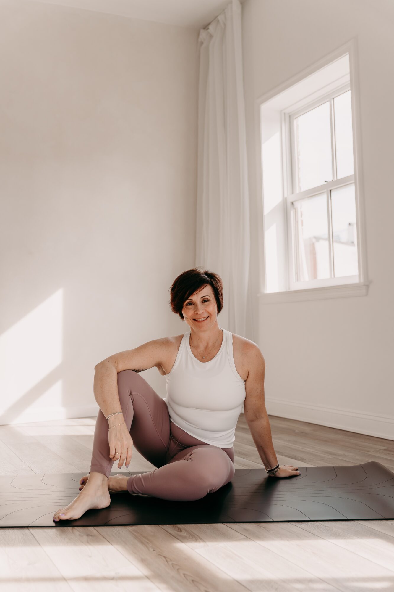 Woman sitting on yoga mat in bright room with large window, smiling, wearing white tank top and leggings.