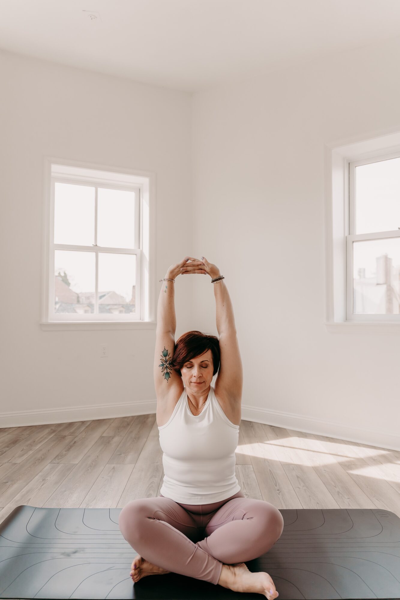 Person sitting cross-legged on yoga mat with arms raised overhead in a bright room with two windows.