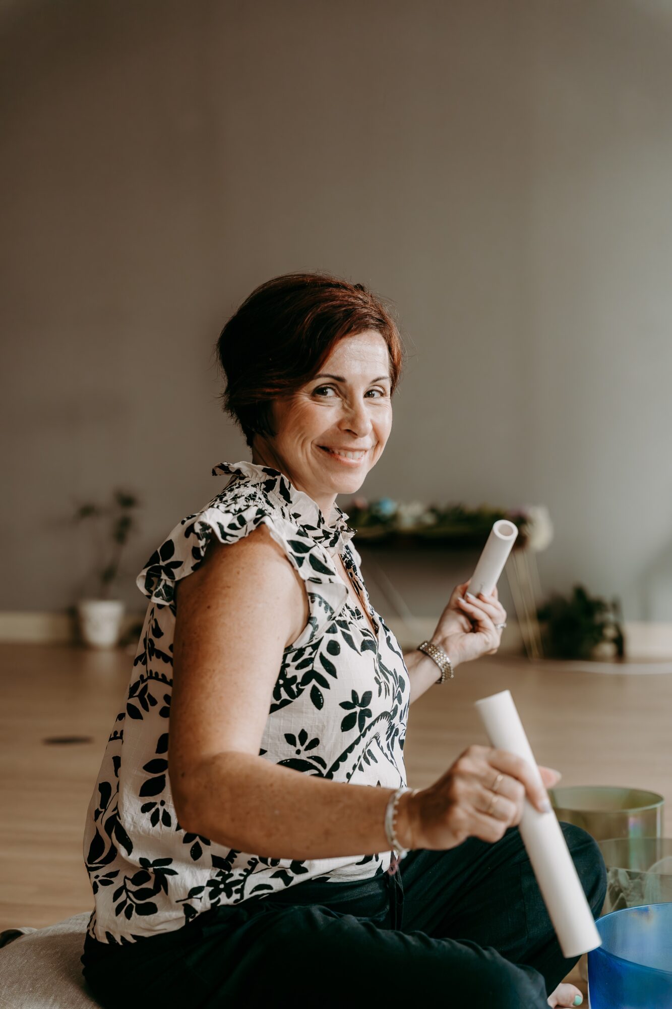 Woman with short dark hair smiling, holding a rolled paper in each hand, wearing a sleeveless floral top, sitting indoors.