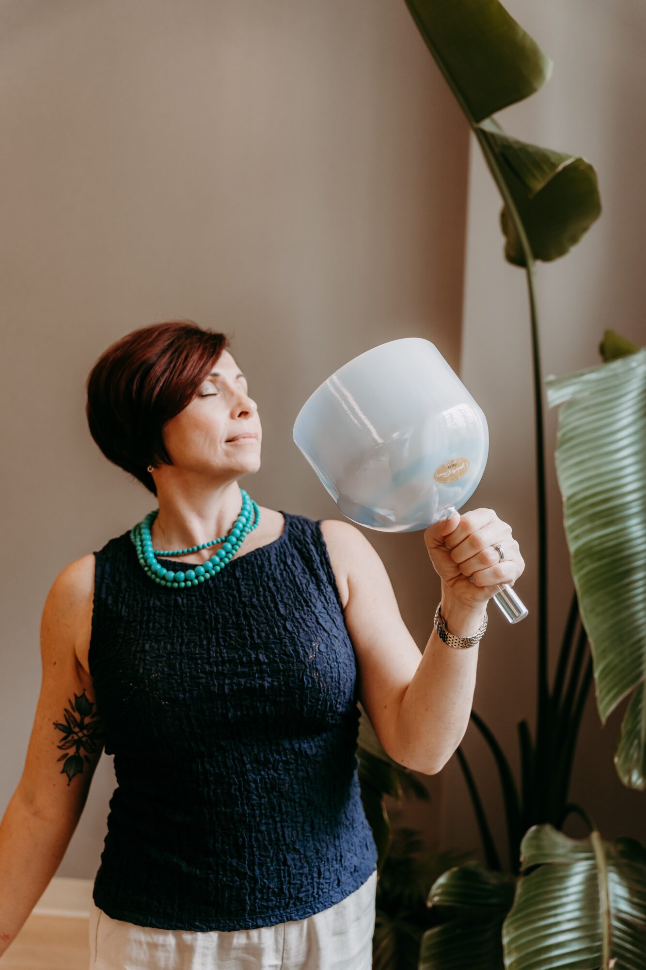 Woman with short hair holding a white bowl, looking to the side, in a room with large green plants.